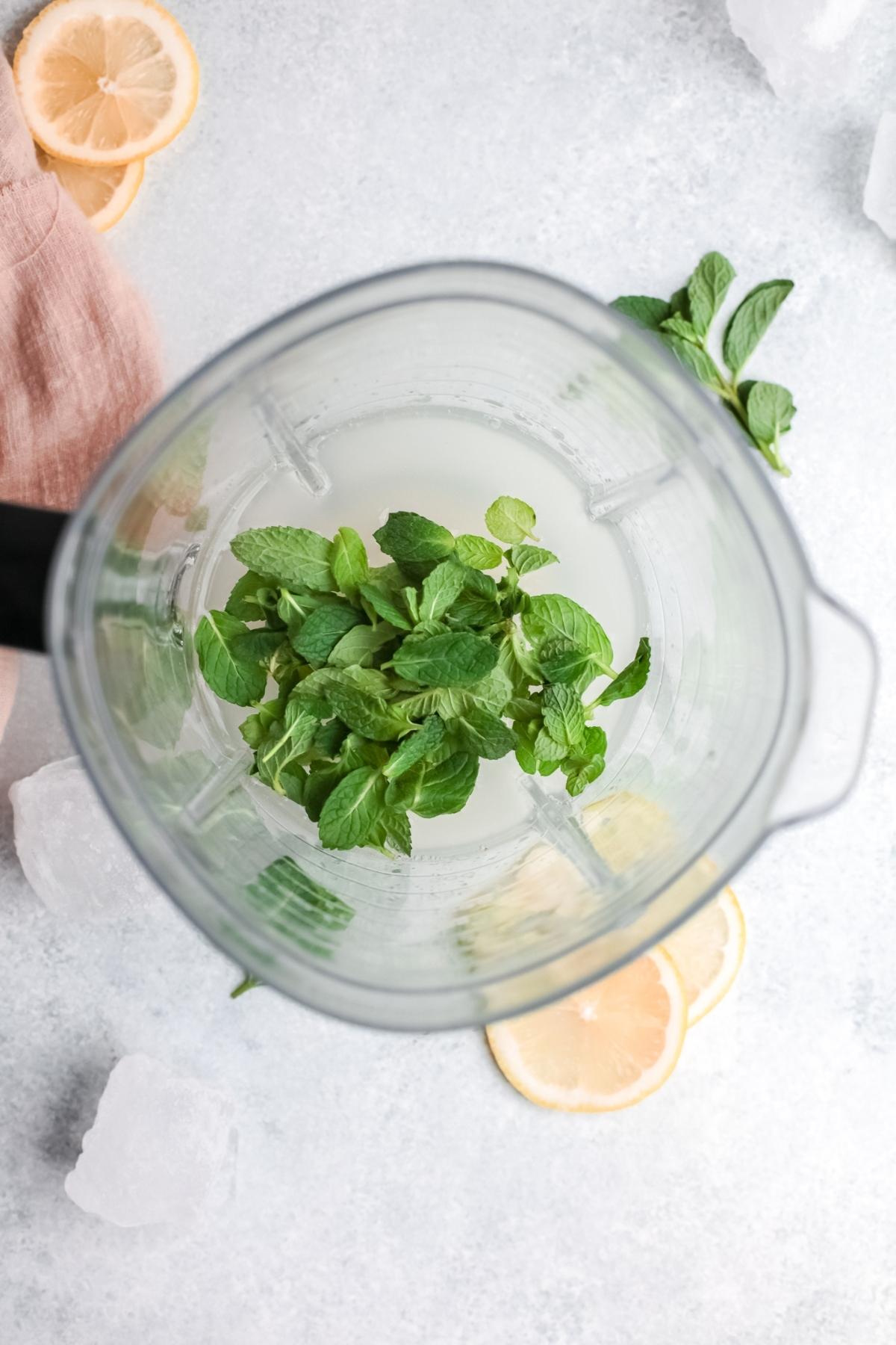 Overhead view of a blender with mint leaves and liquid, surrounded by lemon slices and ice cubes—perfect for making Frozen Mint Lemonade - limonana or trying a refreshing limonana frozen lemonade recipe.