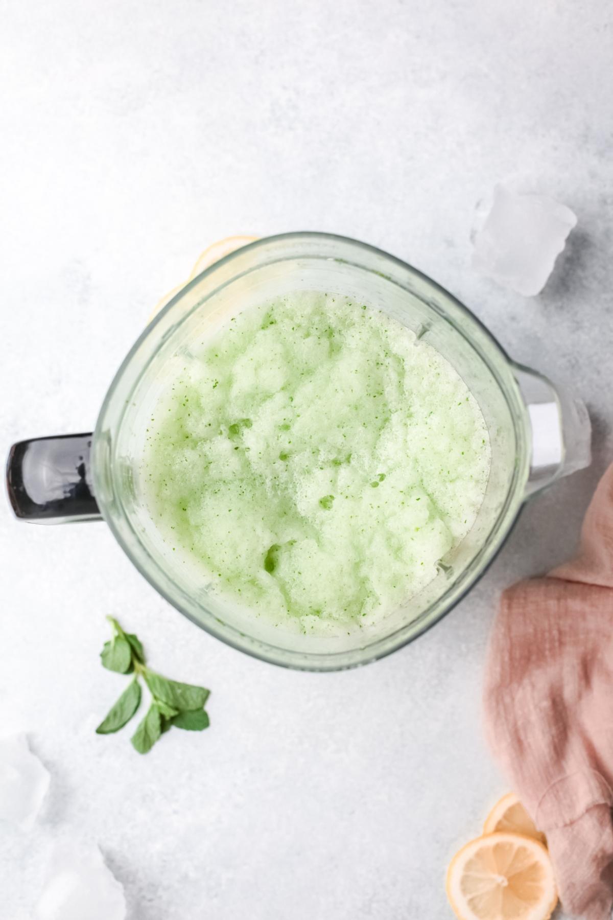 A blender filled with a frothy, light green Frozen Mint Lemonade - limonana, surrounded by mint leaves, lemon slices, and ice cubes.