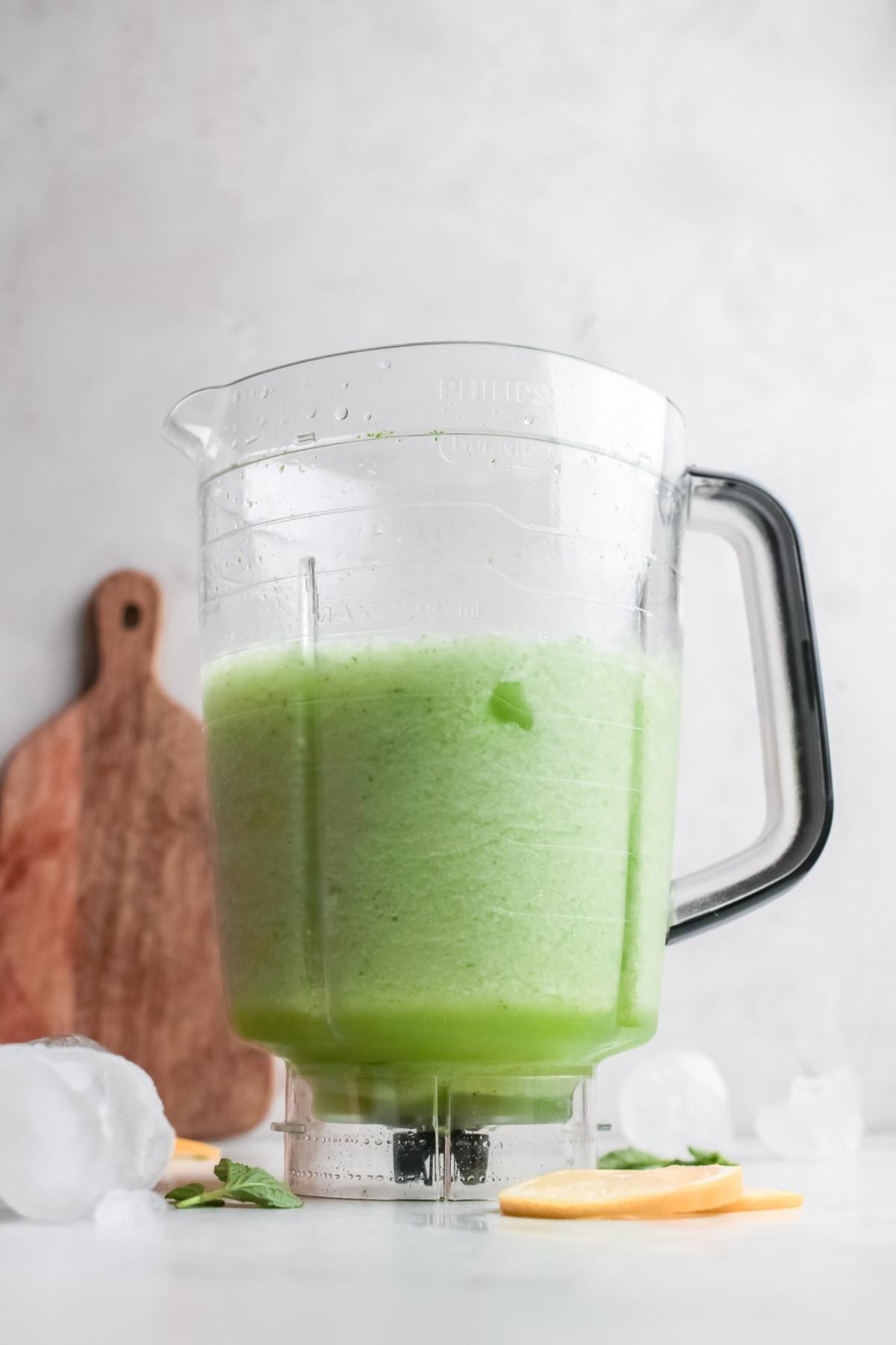 A blender pitcher filled with light green smoothie, reminiscent of a Refreshing Limonana, with ice cubes and lemon slices on the table nearby.