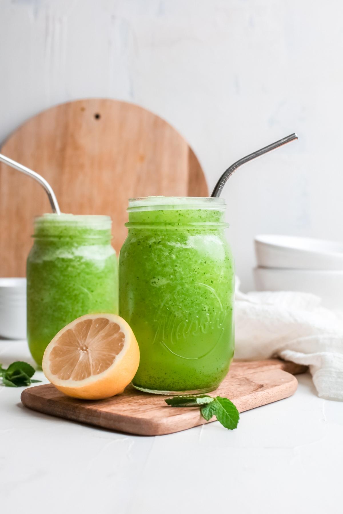 Two mason jars filled with Frozen Mint Lemonade - limonana, metal straws, mint, and a lemon half on a wooden board.