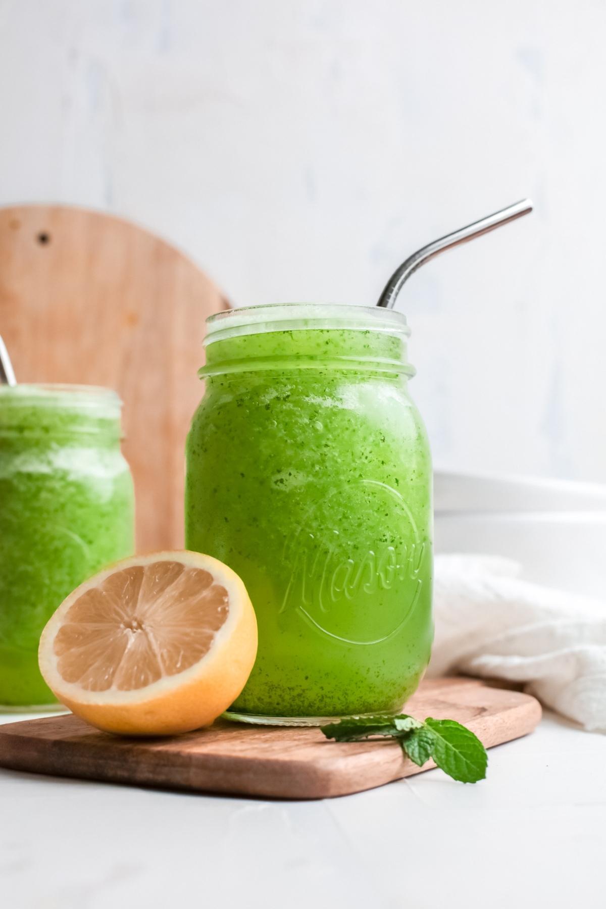 A mason jar of green smoothie with a metal straw sits next to a half lemon and mint leaves on a wooden board, perfect for those who love a classic lemon and mint drink.