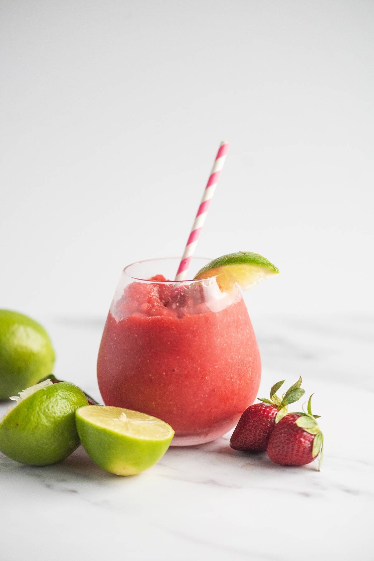 A glass of frozen strawberry lime daiquiri with a lime wedge, striped straw, limes, and strawberries on a marble surface—a refreshing rum cocktail recipe.