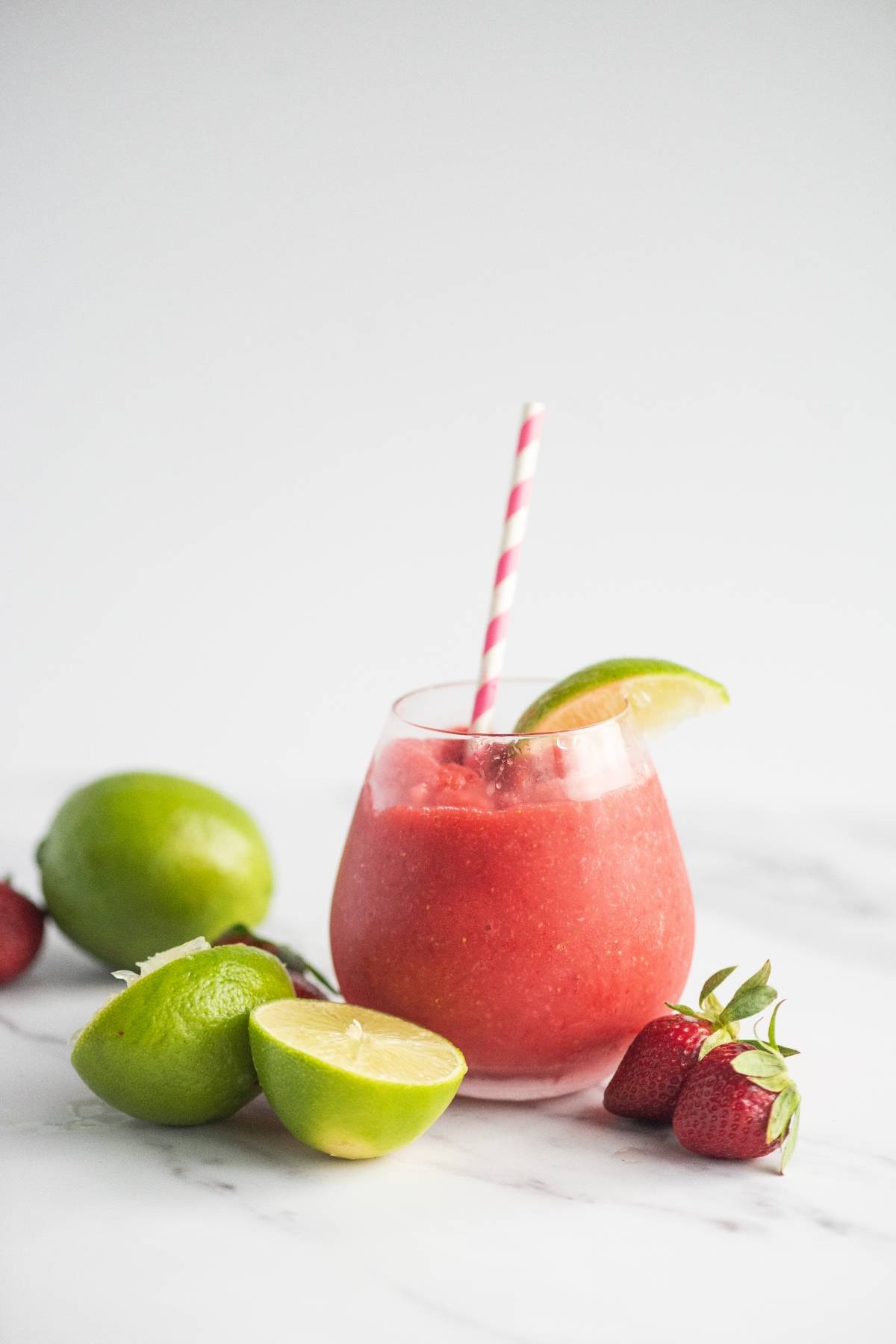A glass of red smoothie with a striped straw, lime wedge, limes, and strawberries on a white surface—an easy way to make a daiquiri or try the best daiquiri recipe for a refreshing twist.