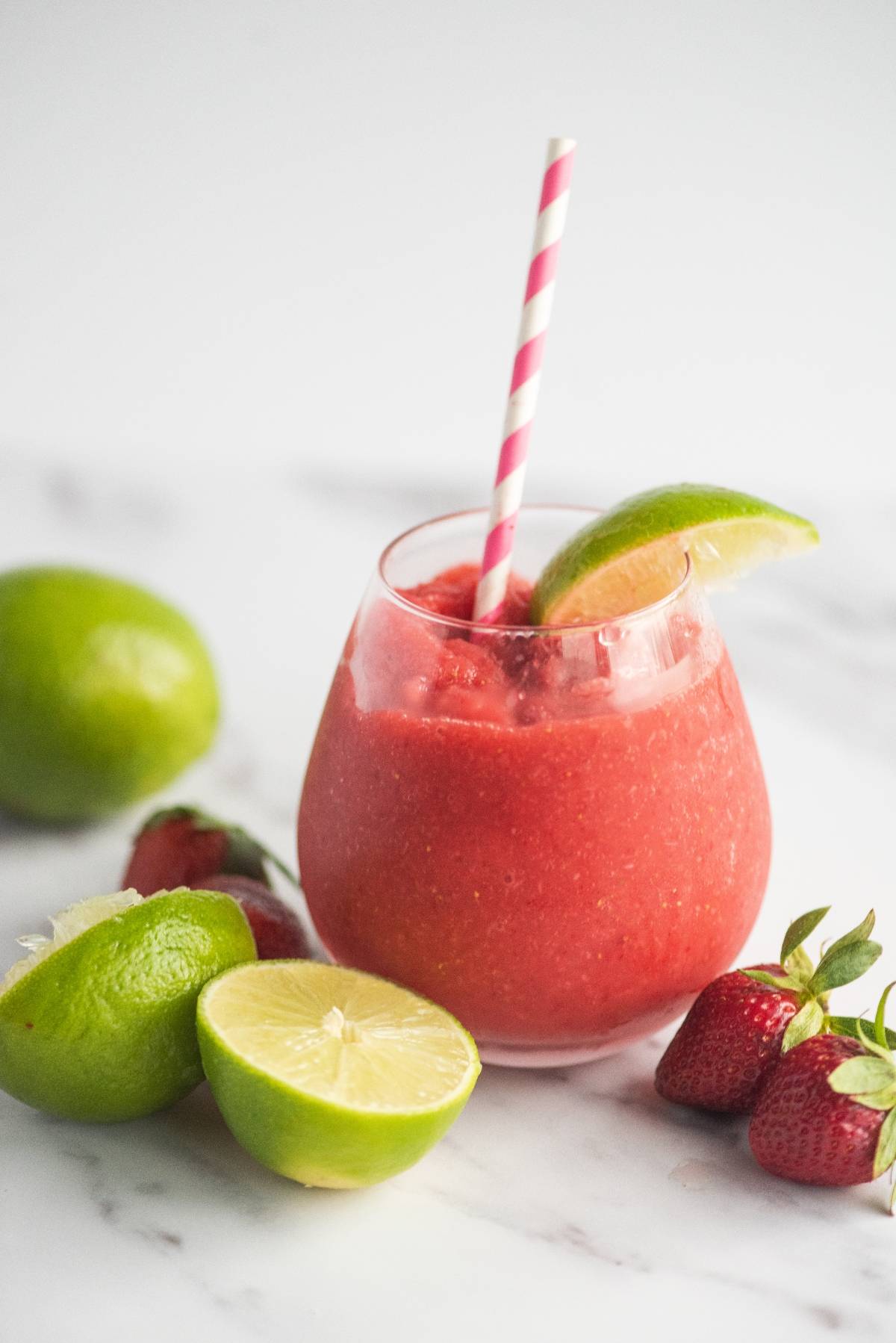 A frozen strawberry lime daiquiri in a glass with a striped straw, garnished with lime, surrounded by limes and strawberries—an easy way to make a refreshing rum cocktail recipe.