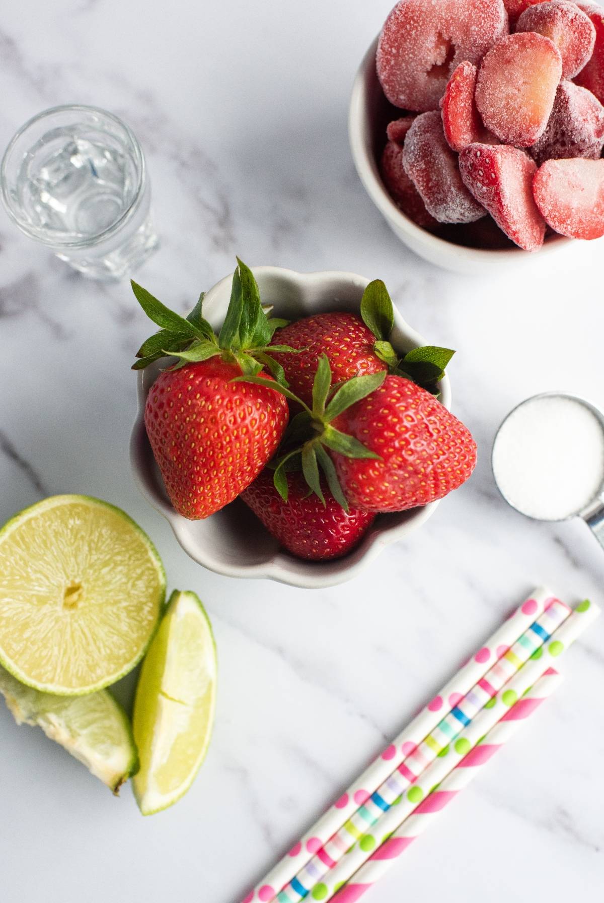 Fresh strawberries, frozen strawberries, lime wedges, sugar, a shot glass, and colorful straws on a white surface—everything you need for the best daiquiri recipe.