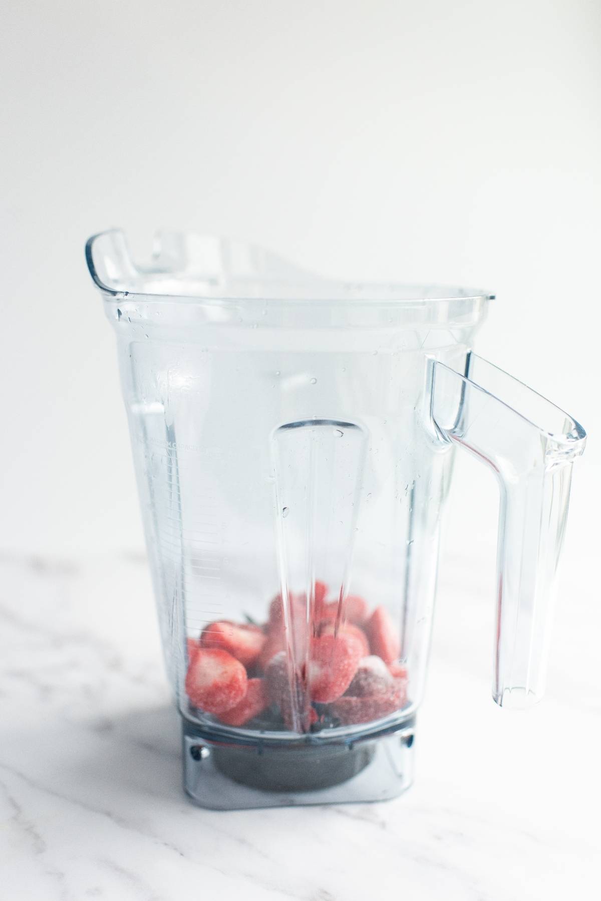 A clear blender pitcher with frozen strawberries inside sits on a white marble countertop, ready to blend into the best daiquiri recipe.