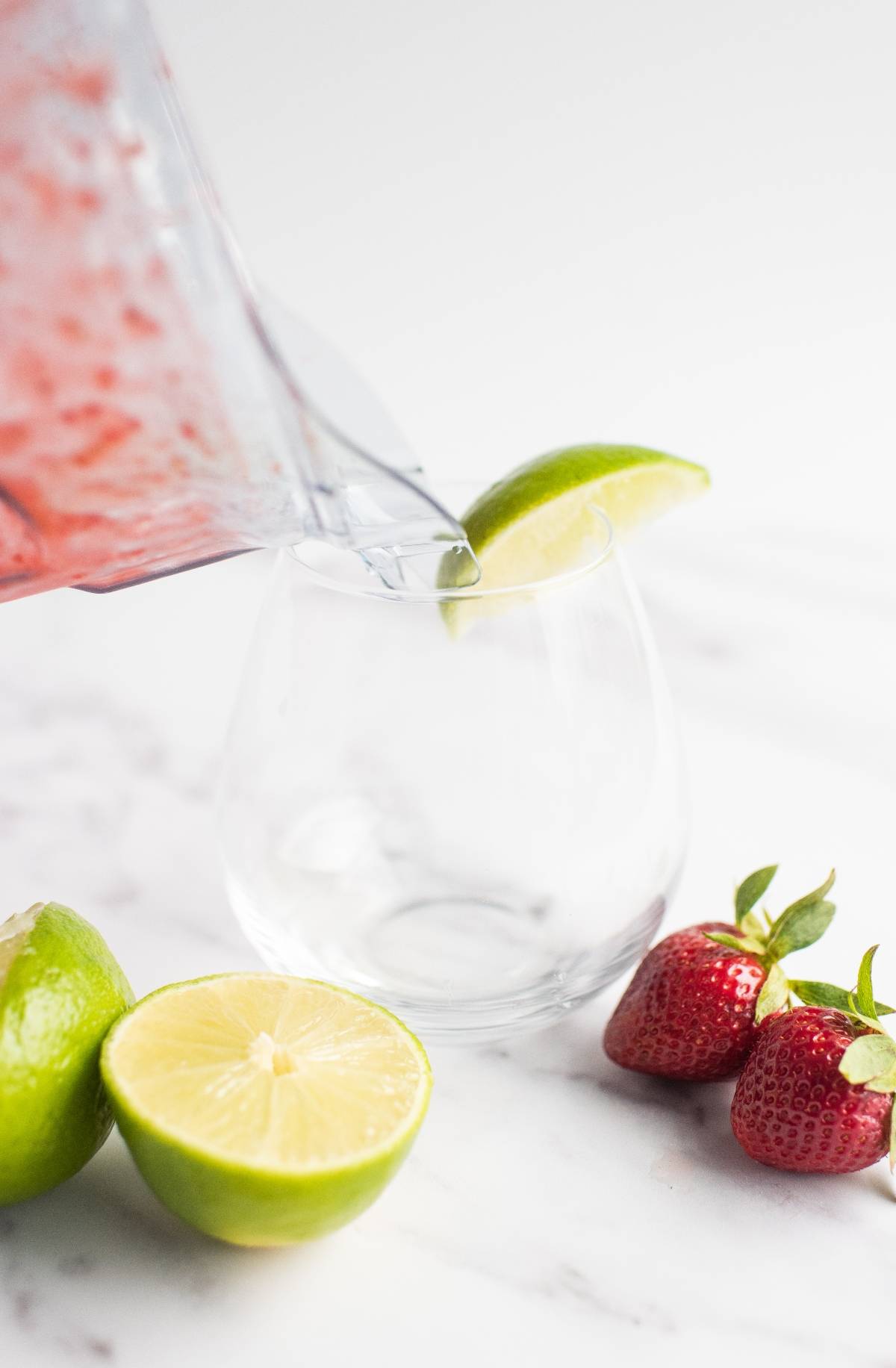 A blender pours a red drink into a glass with a lime wedge; limes and strawberries are nearby, showing the easy way to make a daiquiri or try a frozen strawberry lime daiquiri.