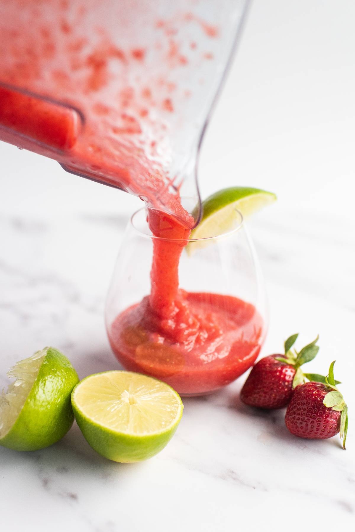 Bright red frozen strawberry lime daiquiri poured into a glass with lime and strawberries on a white surface—the best daiquiri recipe for summer refreshment.