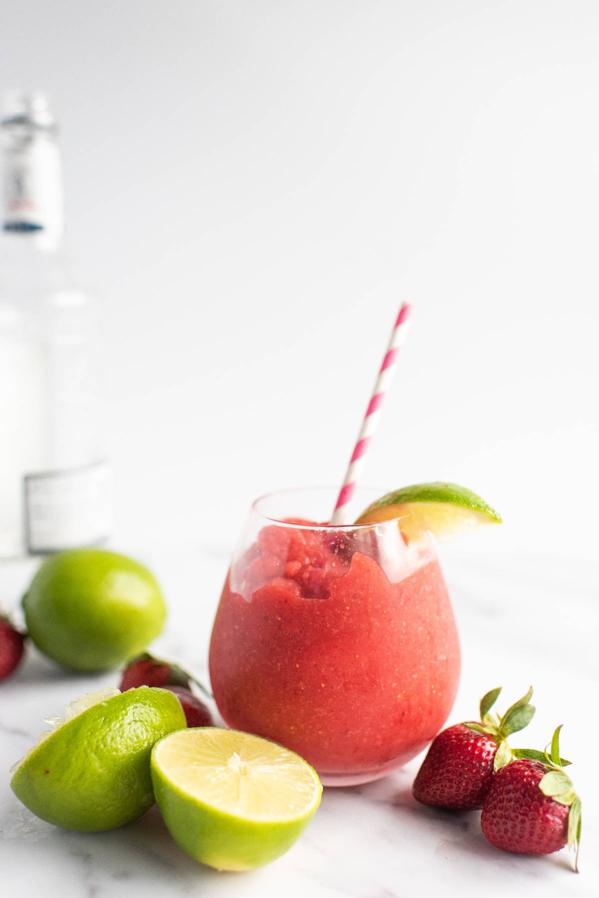 A strawberry slush drink with a lime wedge, surrounded by fresh limes and strawberries, with a striped straw—an easy way to make a daiquiri for the perfect summer rum cocktail recipe.
