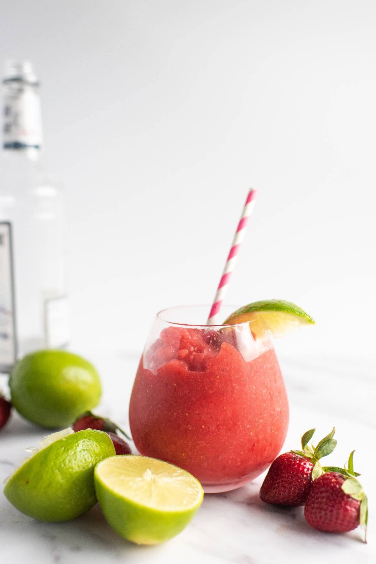 A strawberry frozen drink in a glass with a lime wedge, surrounded by limes and strawberries on a white surface—perfect for trying the best daiquiri recipe or enjoying a refreshing frozen strawberry lime daiquiri.