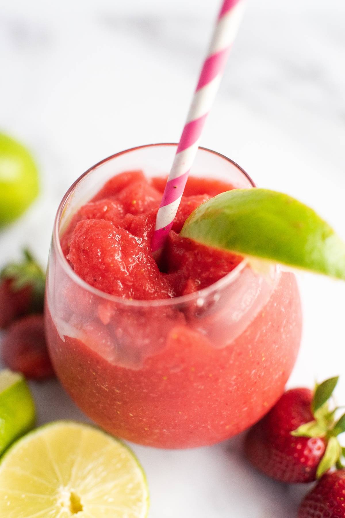A glass of frozen strawberry lime daiquiri with a pink straw and a lime wedge, surrounded by limes and strawberries—a refreshing rum cocktail recipe perfect for summer.