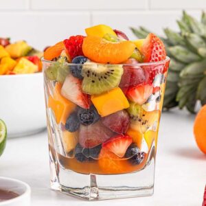 Glass of honey lime fruit salad with strawberries, kiwi, blueberries, grapes, and mango, surrounded by fresh fruits.