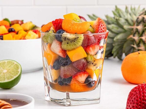 Glass of honey lime fruit salad with strawberries, kiwi, blueberries, grapes, and mango, surrounded by fresh fruits.
