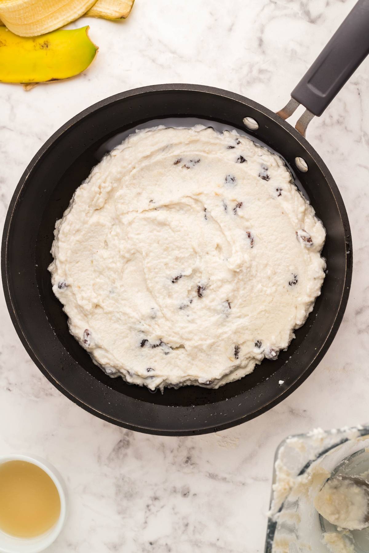 A skillet filled with Gluten Free Kaiserschmarrn batter, an Austrian and German dessert pancake dotted with raisins, sits on a marble countertop.