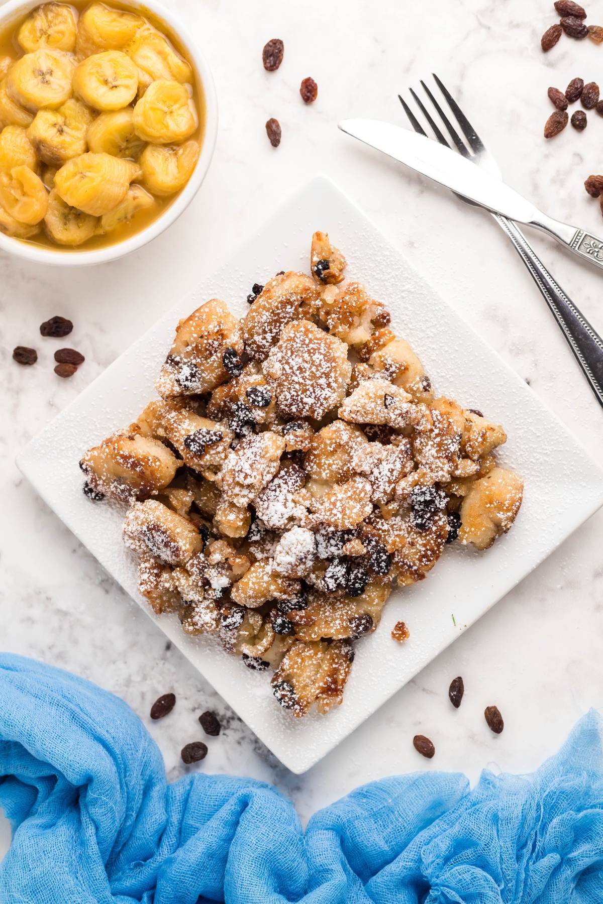 A plate of powdered sugar-dusted Austrian and German dessert pancakes, known as Kaiserschmarrn with raisins, sits next to a bowl of cooked bananas.