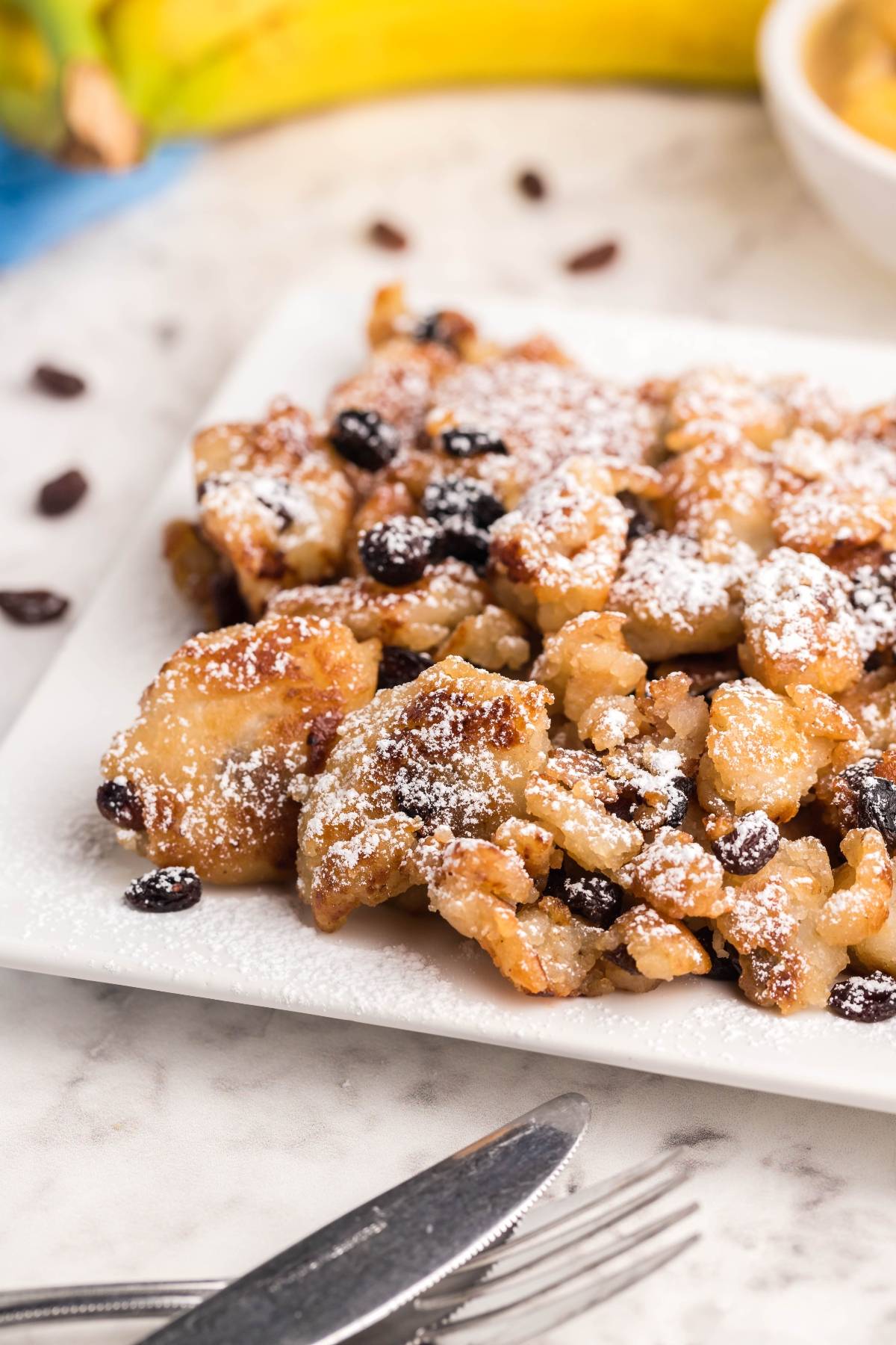 Plate of Gluten Free Kaiserschmarrn with raisins, dusted with powdered sugar, on a marble table next to utensils—a delightful gluten free dessert pancake experience.