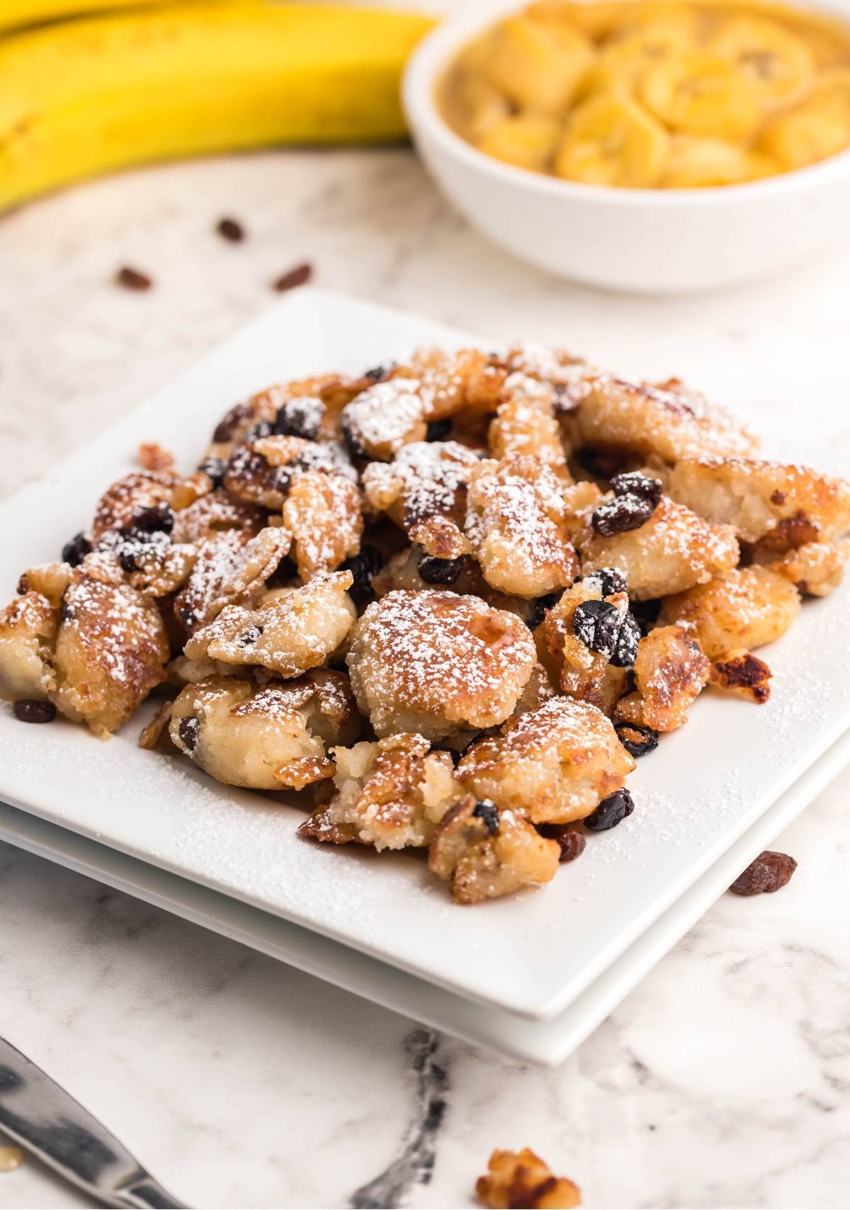 A plate of powdered sugar-dusted banana fritters with raisins on a white square dish, inspired by Austrian and German dessert pancakes.