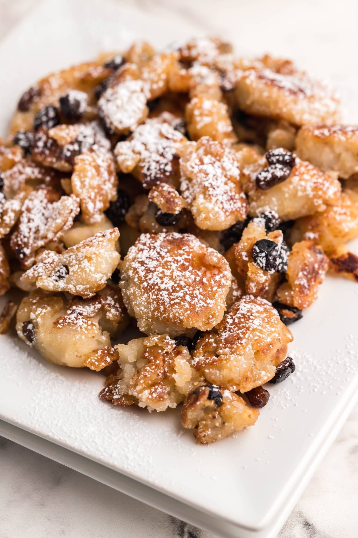 A plate of golden, powdered sugar-dusted Gluten Free Kaiserschmarrn with raisins on a white square dish—an irresistible Austrian and German dessert pancake, perfectly scrambled and sweet.