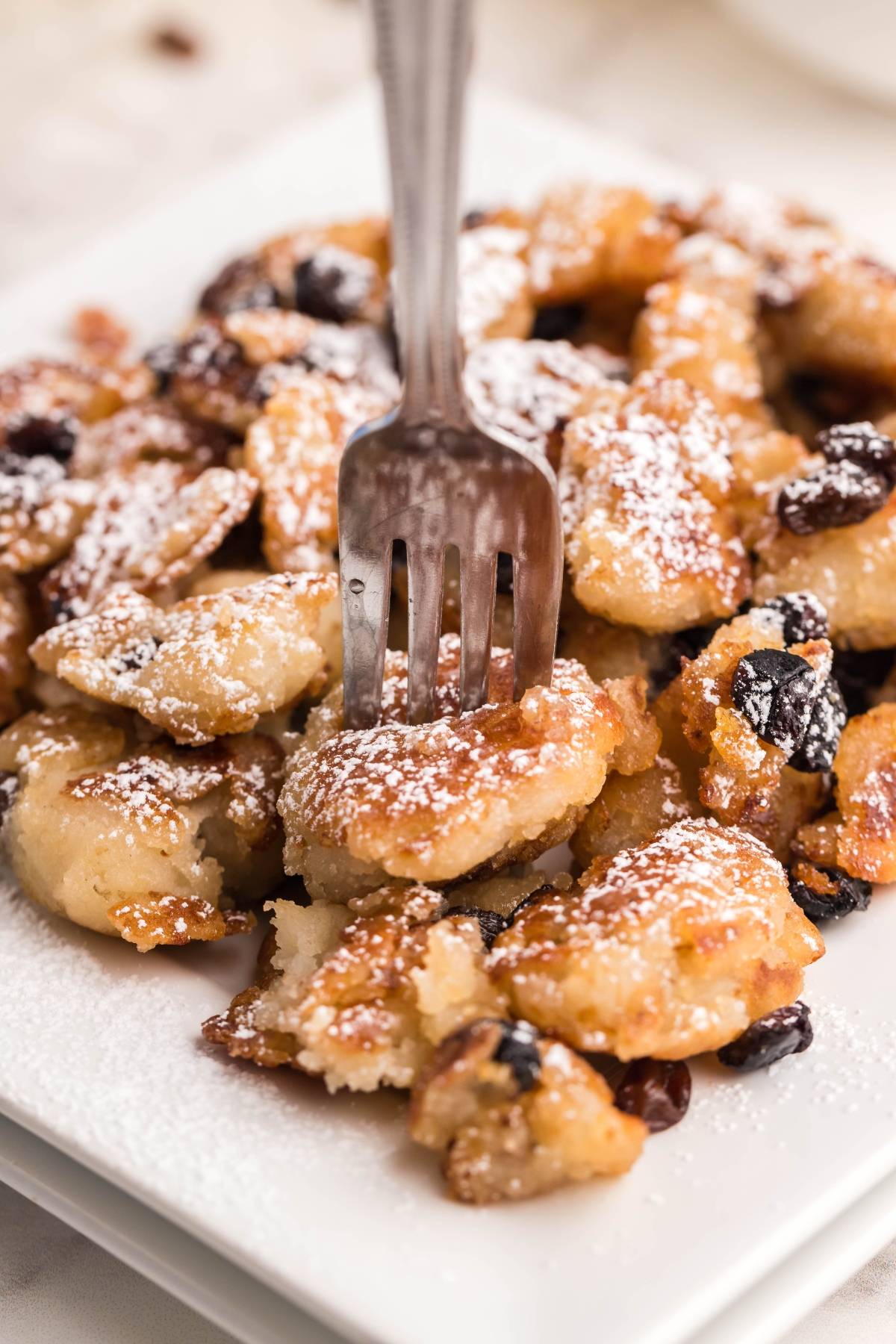 A fork in fluffy, golden Kaiserschmarrn pancake pieces with raisins, dusted with powdered sugar on a white plate—an irresistible Austrian and German dessert pancake.