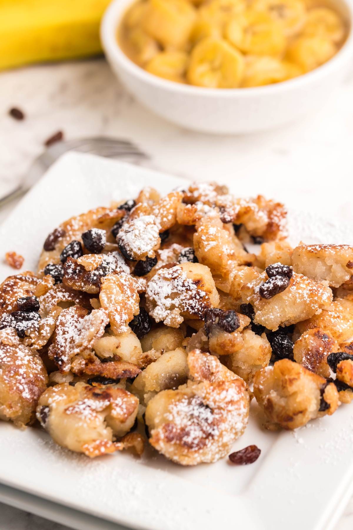 A plate of Kaiserschmarrn, the classic Austrian and German dessert pancake, topped with powdered sugar and raisins, with a bowl of sliced bananas in the background.