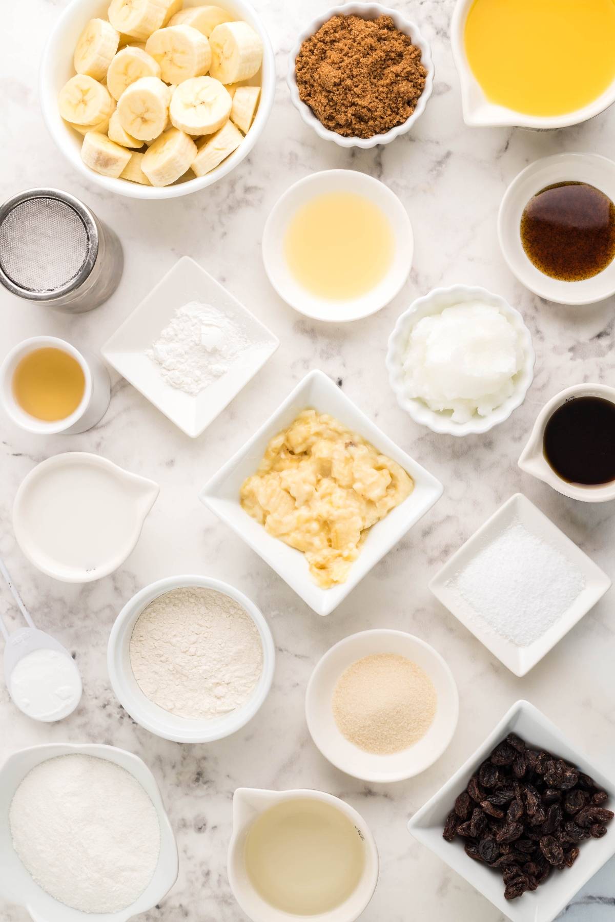 Assorted baking ingredients in bowls, perfect for making gluten free dessert pancakes like Gluten Free Kaiserschmarrn, including bananas, flour, oil, raisins, sugar, and various liquids on a marble surface.