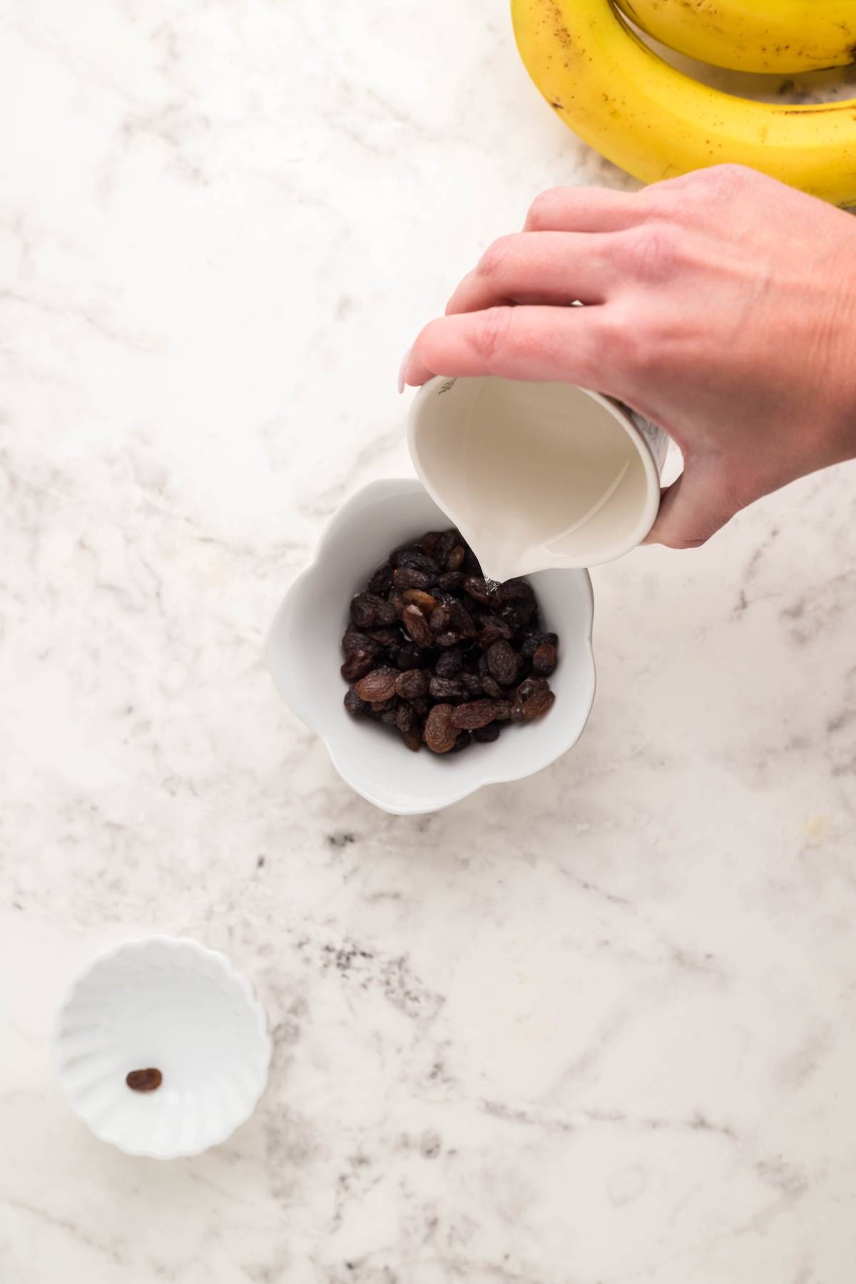 A hand pours liquid from a small pitcher into a bowl of raisins on a marble countertop, preparing ingredients for a gluten free dessert pancake.