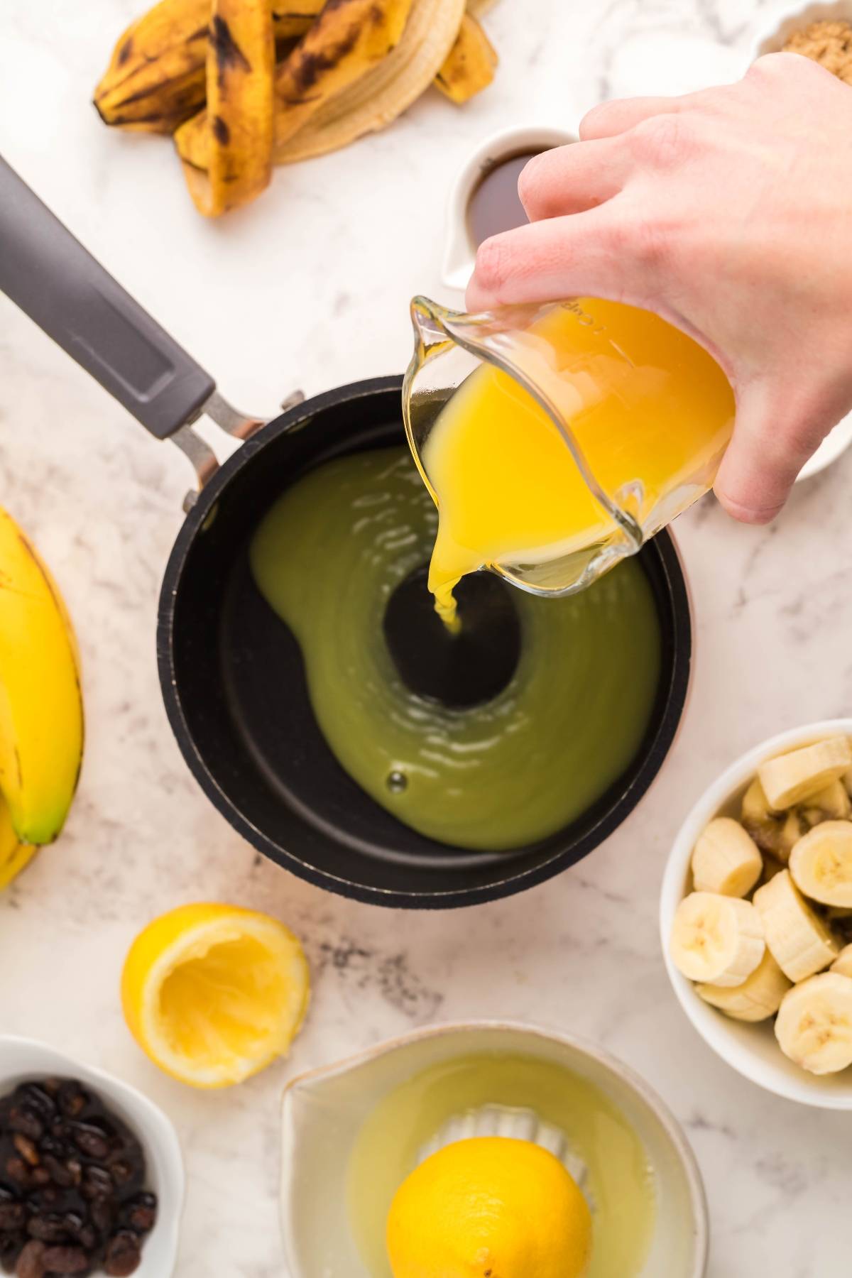 A hand pours orange juice into a saucepan surrounded by bananas, lemon, and other ingredients on a white surface—perfect steps for how to make Kaiserschmarrn, the beloved Austrian and German dessert pancakes.