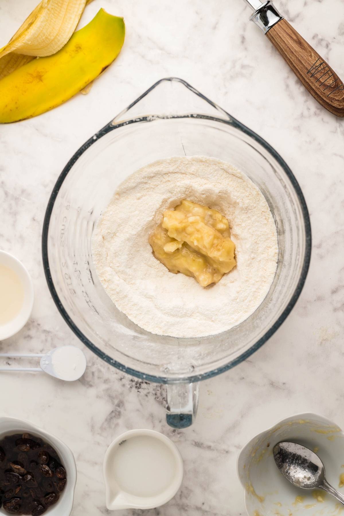 Overhead view of a blender with flour and mashed banana, surrounded by baking ingredients on a marble counter—perfect for making Gluten Free Kaiserschmarrn, the classic scrambled pancakes turned into a gluten free dessert pancake delight.
