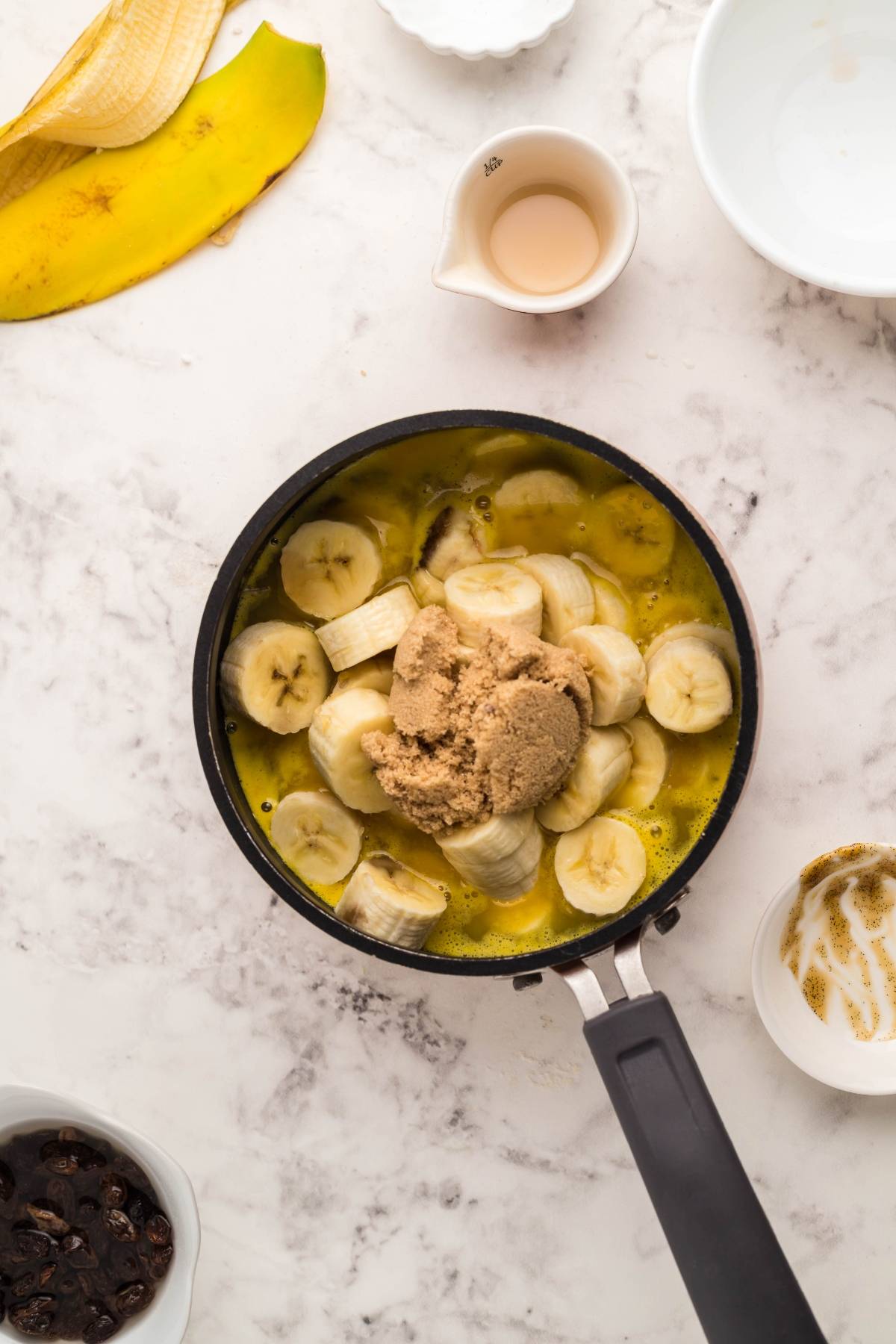 Sliced bananas and brown sugar in a pan with melted butter on a marble countertop, surrounded by bowls—perfect toppings for Gluten Free Kaiserschmarrn, the beloved Austrian and German dessert pancake.