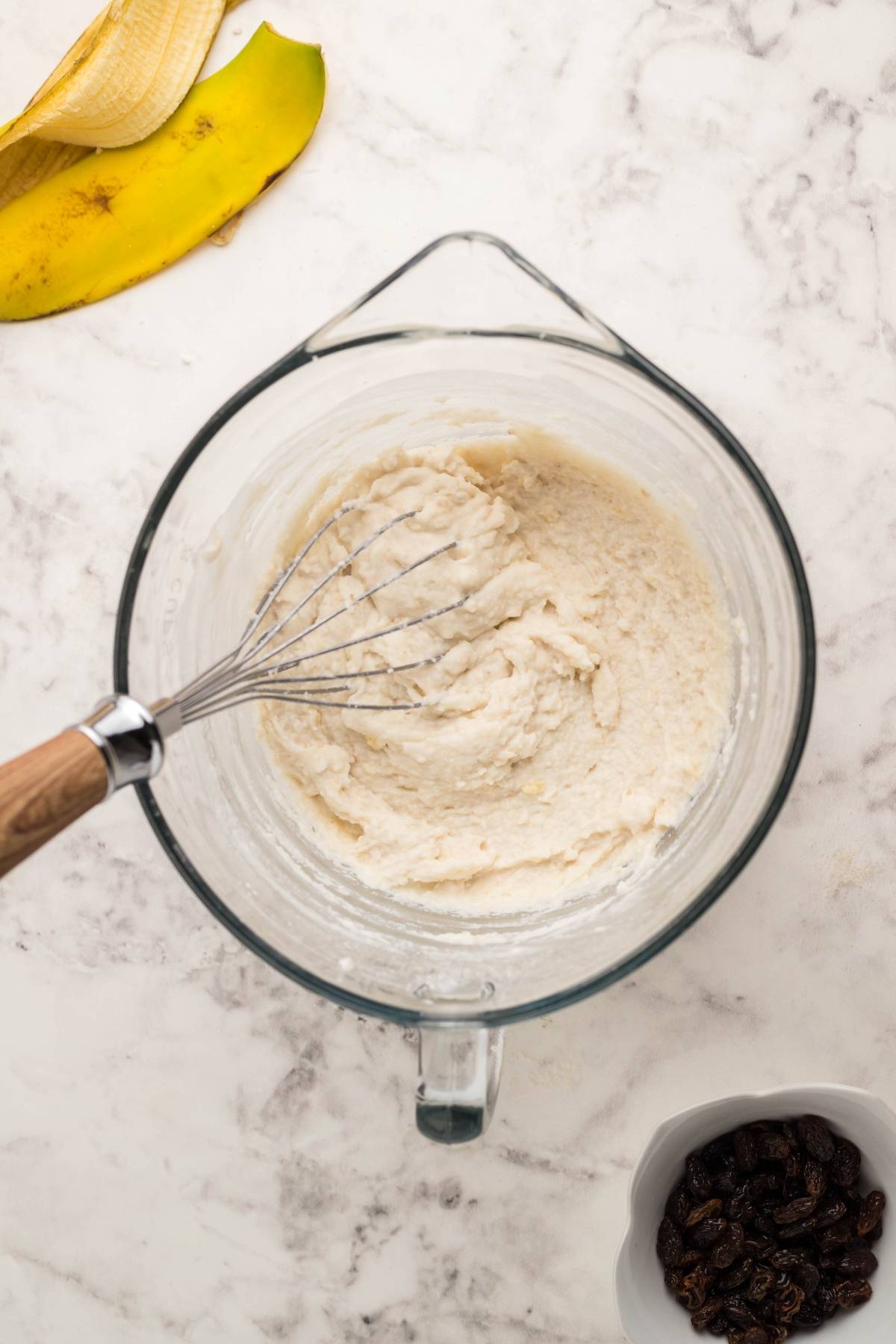 Glass bowl with banana bread batter and a whisk, next to a banana peel and a small bowl of raisins—perfect for making Gluten Free Kaiserschmarrn or other gluten free dessert pancake recipes.
