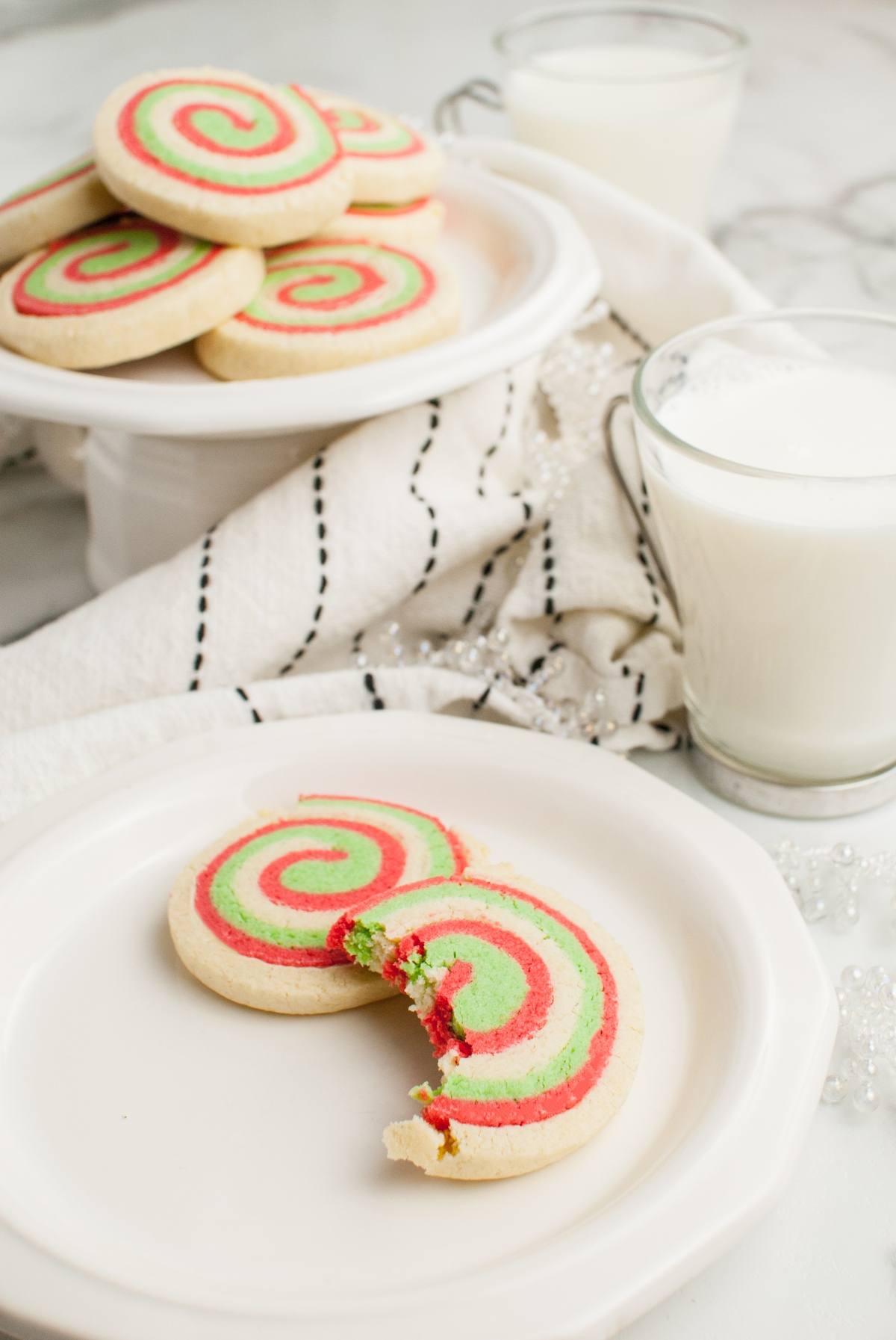 A bitten red, green, and white christmas cookie roll sits on a plate with more red and green Christmas cookies and glasses of milk nearby—perfect for gluten free holiday baking.