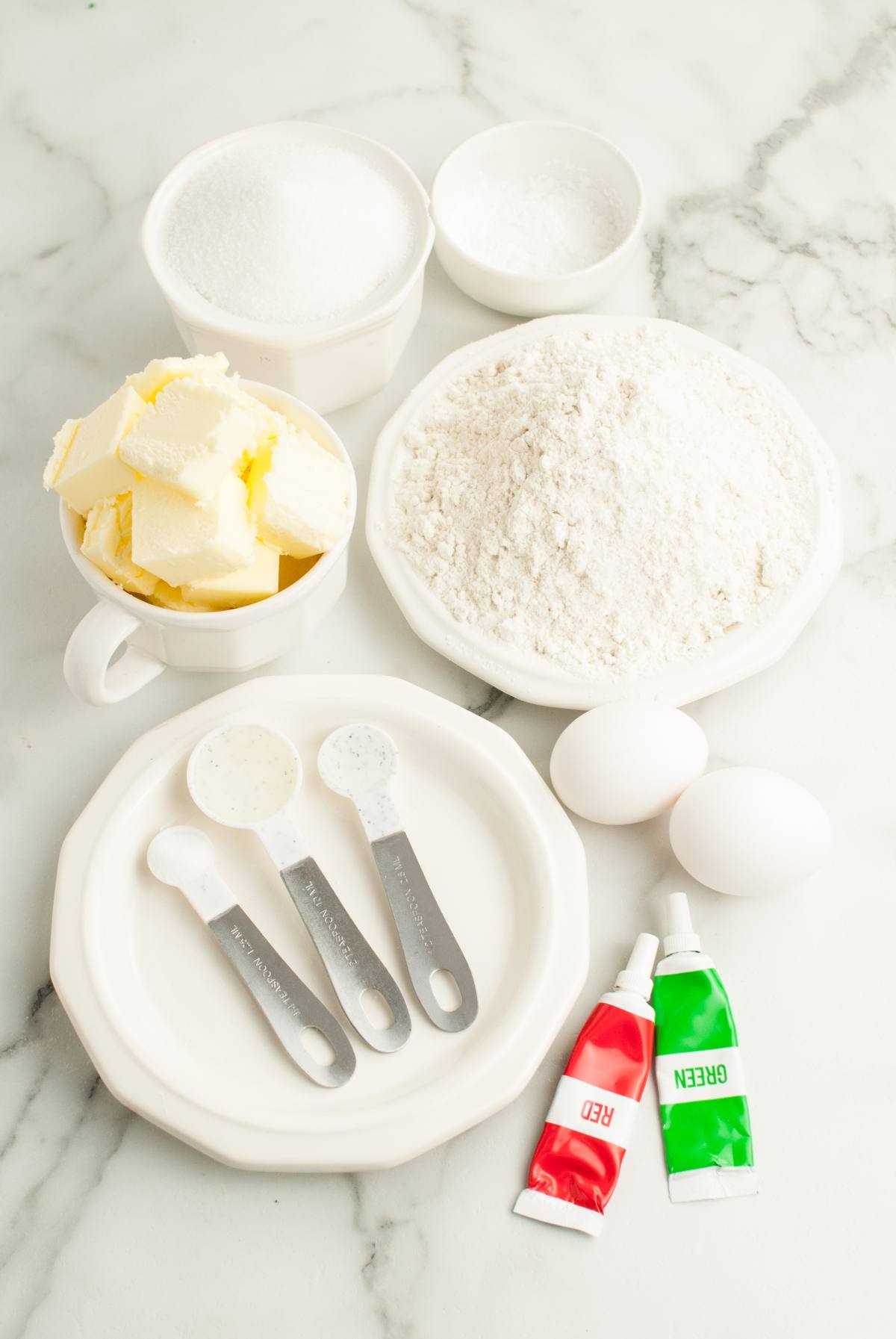 Baking ingredients on a marble surface: flour, sugar, butter, eggs, food coloring, and measuring spoons—perfect for creating festive red and green Christmas cookies like Italian Christmas swirl cookies or Gluten Free Pinwheel Cookies.