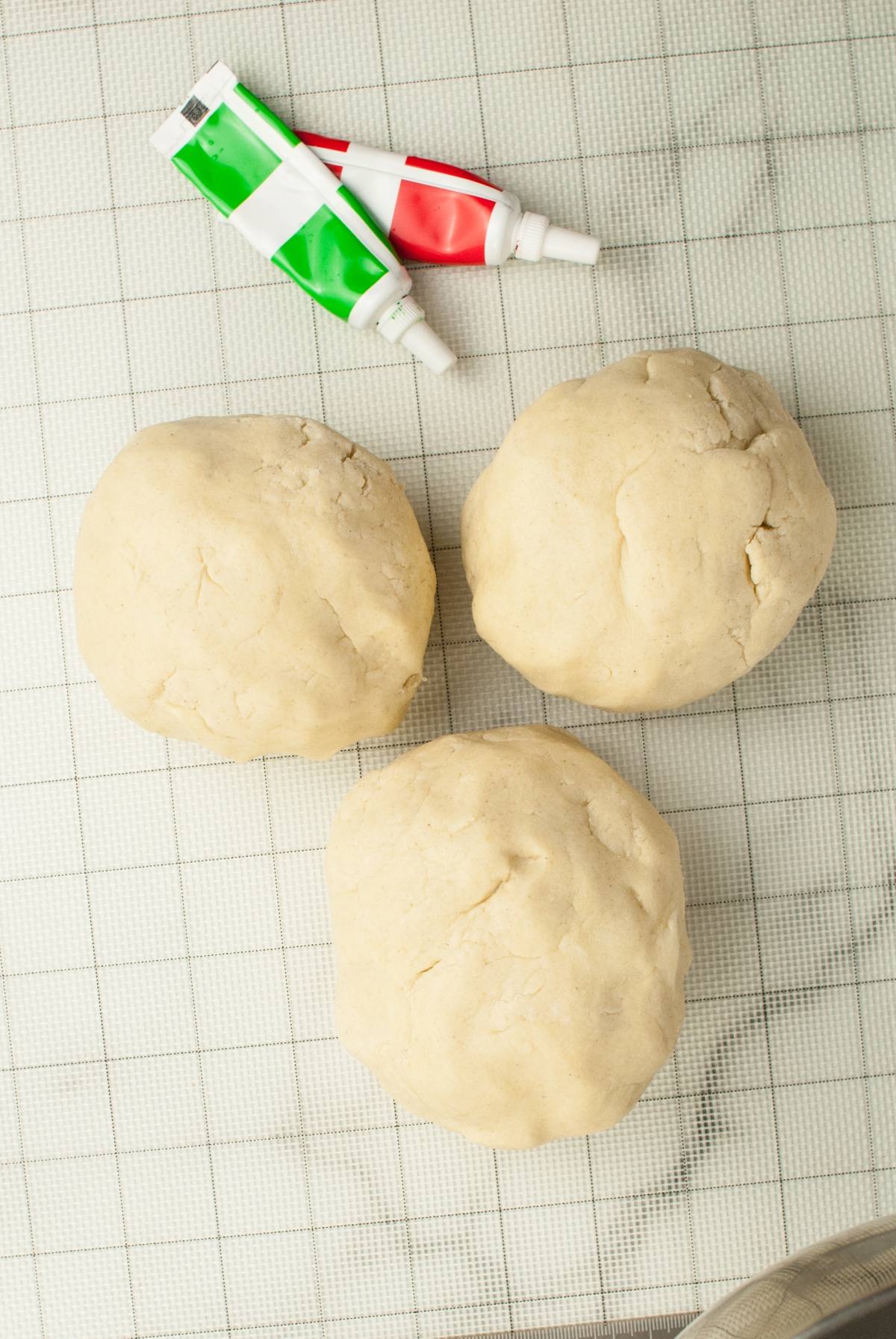 Three balls of dough on a grid surface with red and green food coloring tubes above them, perfect for making festive Italian Christmas swirl cookies.