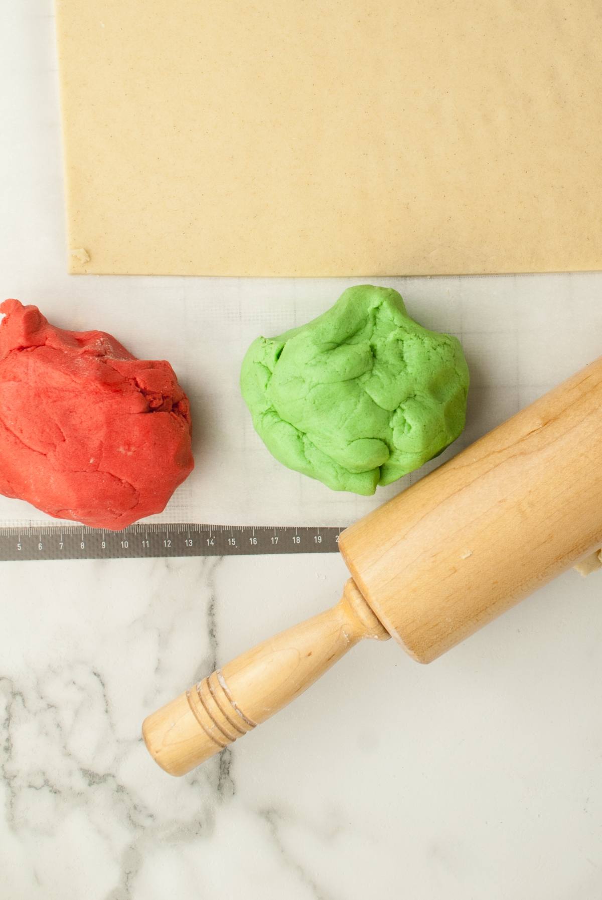 Red and green dough balls, a rolling pin, and a sheet of dough on a marble surface ready to make festive Italian christmas swirl cookies.