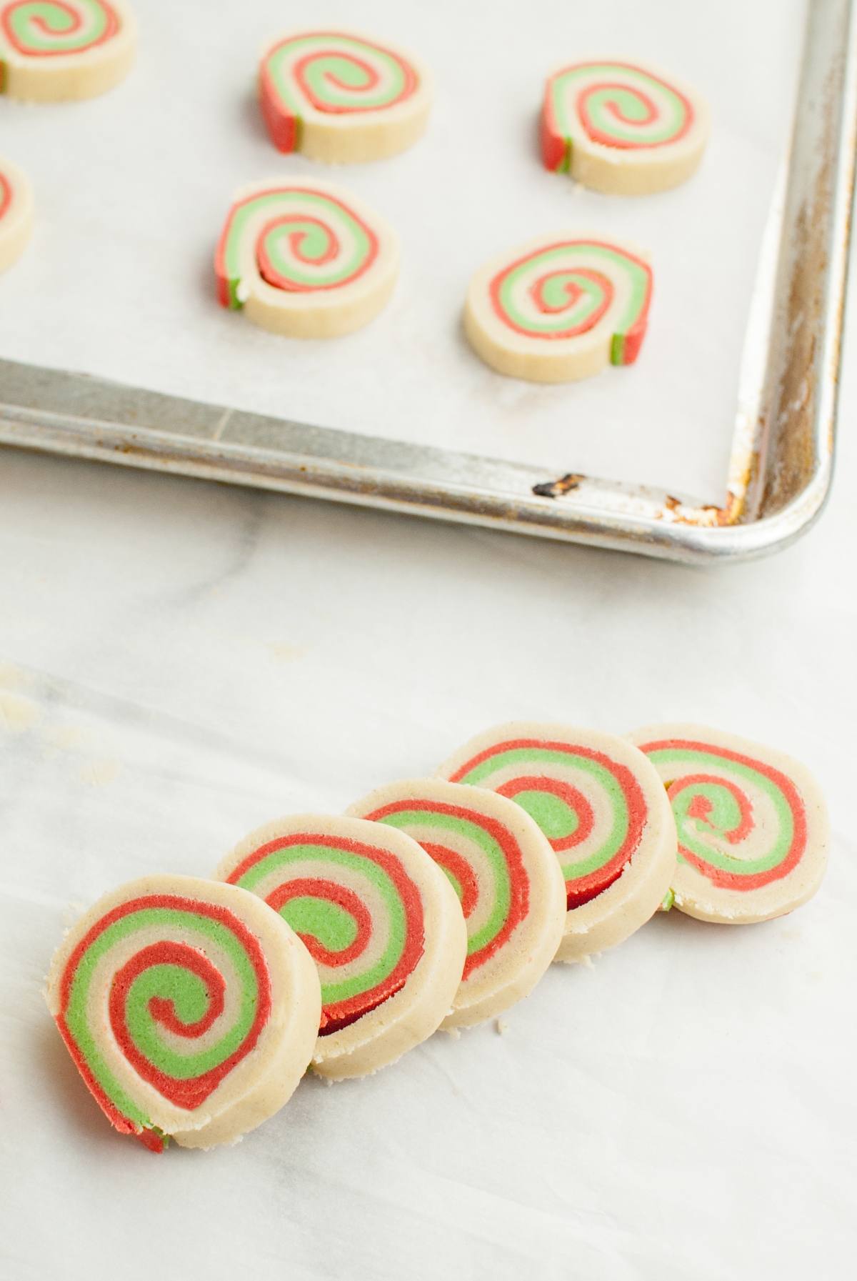Five festive red and green Gluten Free Pinwheel Cookies rest on the counter, with more Christmas cookie rolls baking on a sheet in the background.
