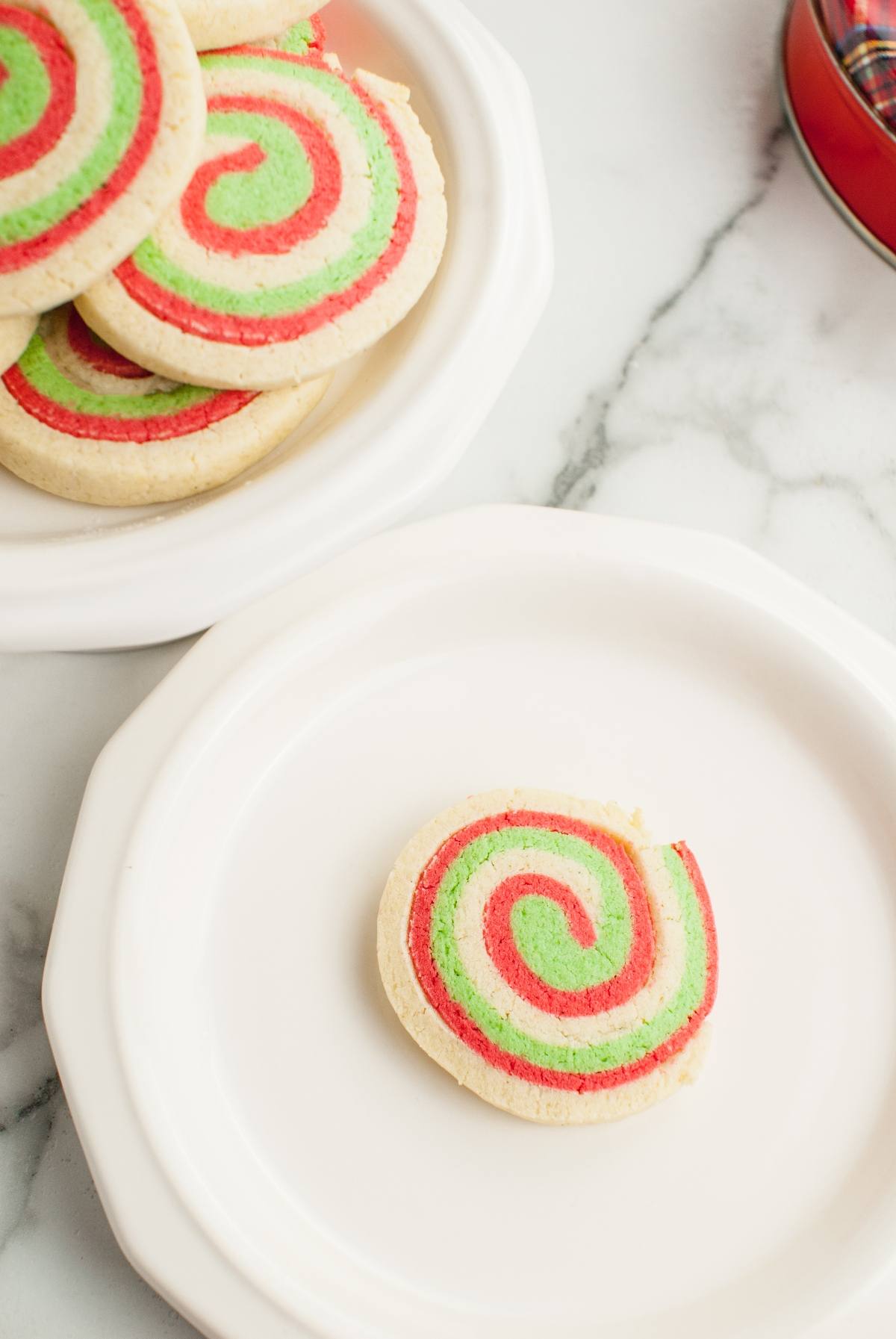 A red, green, and white Italian Christmas swirl cookie sits on a white plate, with more festive red and green Christmas cookies on a plate above it—perfect for gluten free holiday baking.