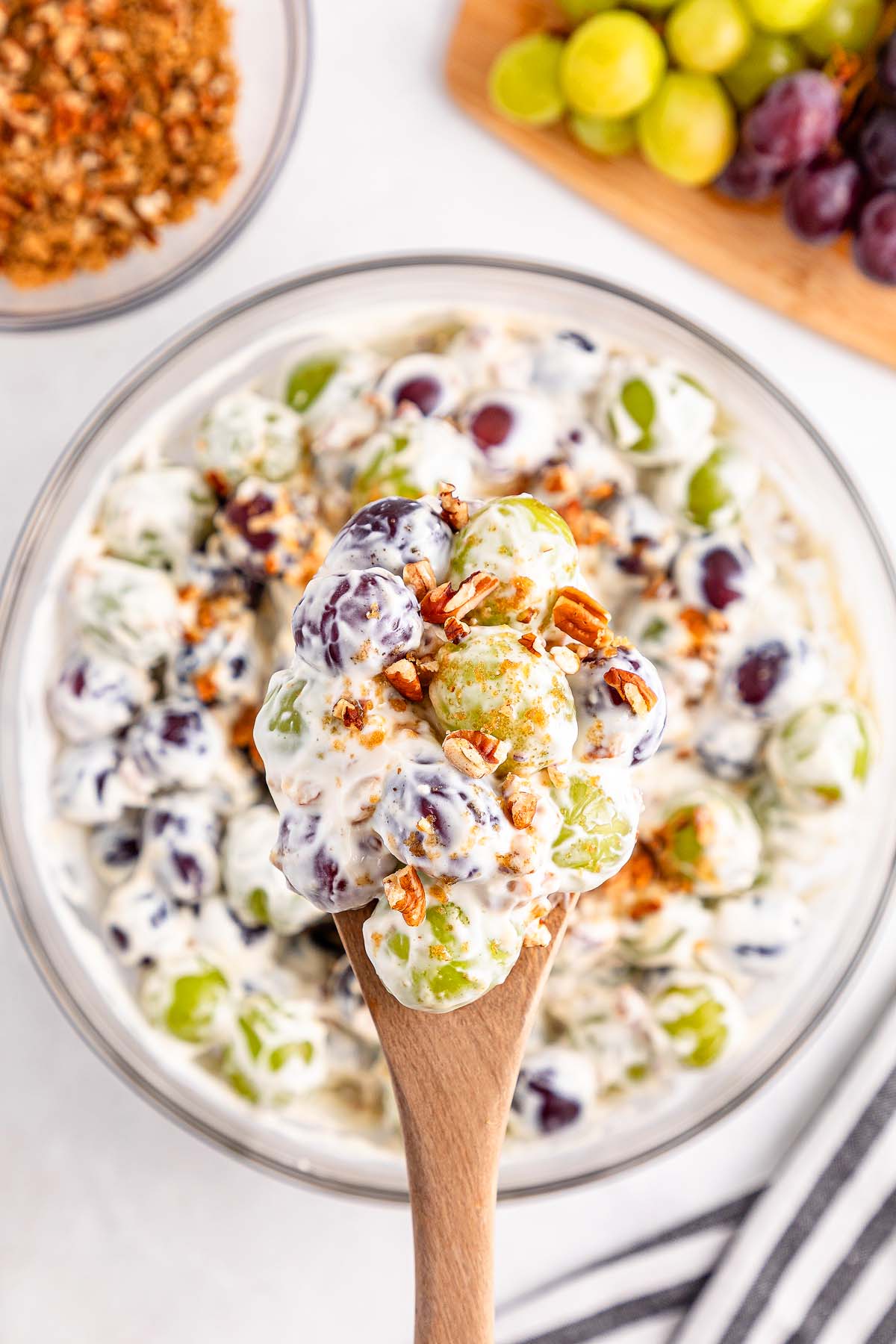 A wooden spoon holds creamy grape salad with granola, poised above a bowl brimming with fresh grape salad.