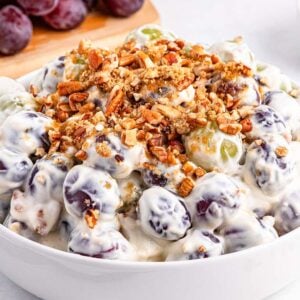 A bowl of creamy grape salad topped with chopped pecans, with fresh grapes and nuts in the background.