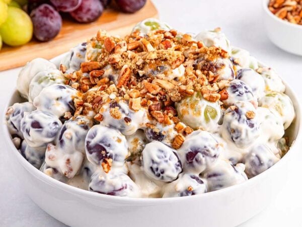 A bowl of creamy grape salad topped with chopped pecans, with fresh grapes and nuts in the background.