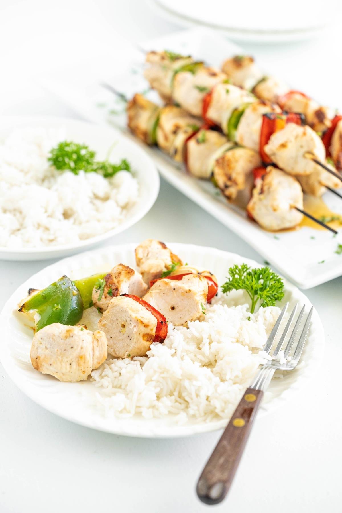 White plate with grilled pepper and chicken kebabs, white rice, and parsley garnish on a table.