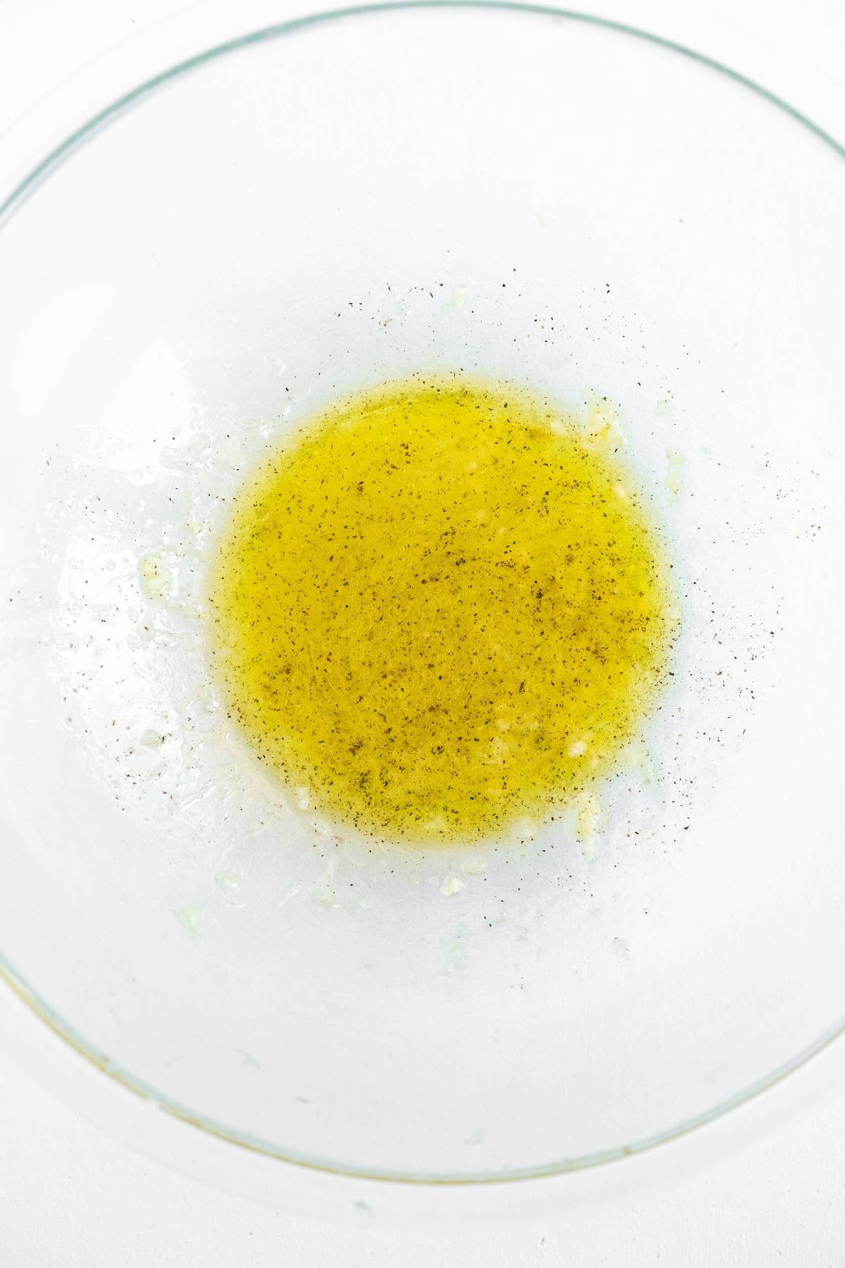 Clear bowl with yellow liquid, speckled with black pepper and seasoning, perfect for marinating tasty chicken skewers or kebabs with chicken and vegetables for your summer BBQ dinner. Set on a white surface.