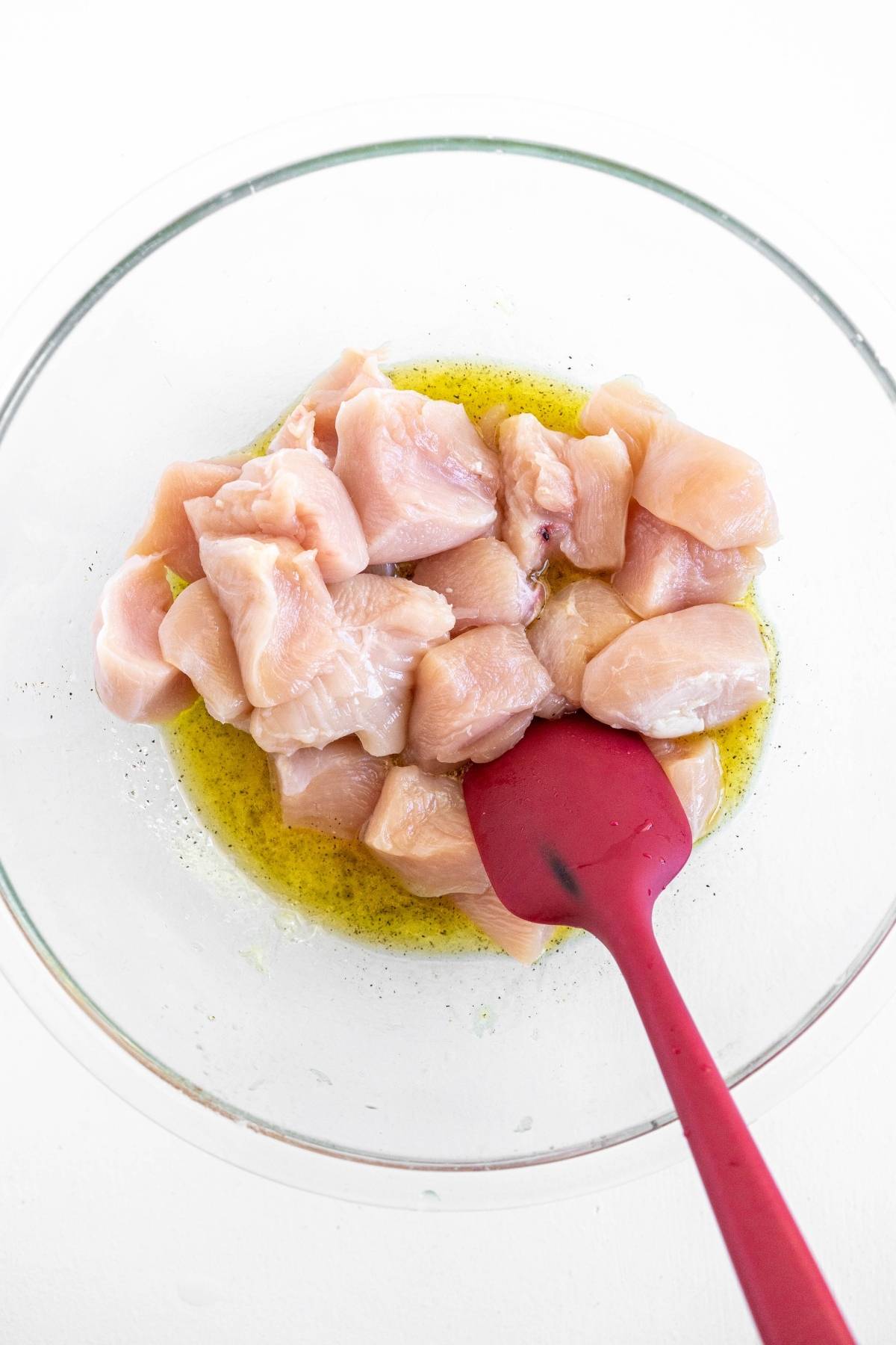 Cubed raw chicken in a glass bowl with marinade, stirred with a red spatula on a white surface—perfect for prepping tasty chicken skewers at your next Summer BBQ dinner.
