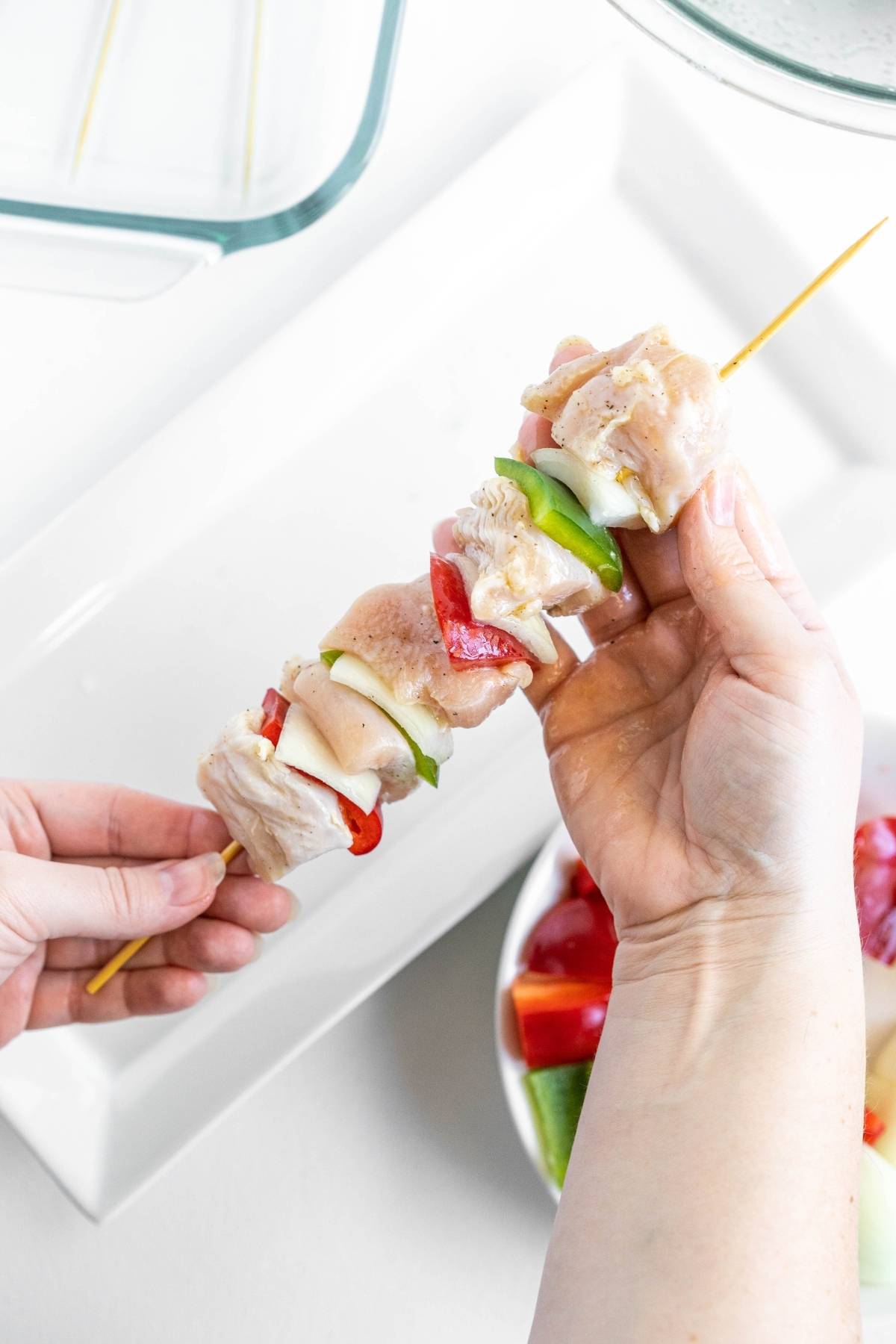 Hands holding a skewer with raw chicken, green pepper, and red pepper pieces over a white plate, perfect for making grilled pepper and chicken kebabs.