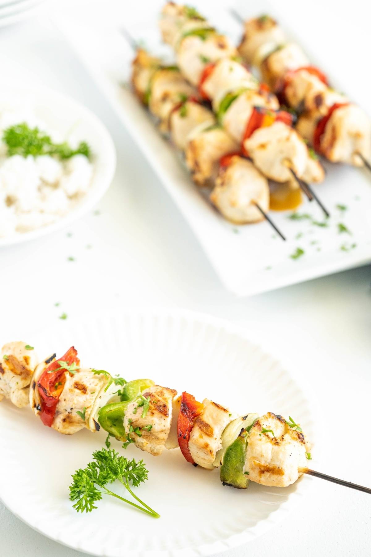 Grilled chicken and vegetable skewer on a white plate, perfect for a summer BBQ dinner, with more skewers and parsley in the background.
