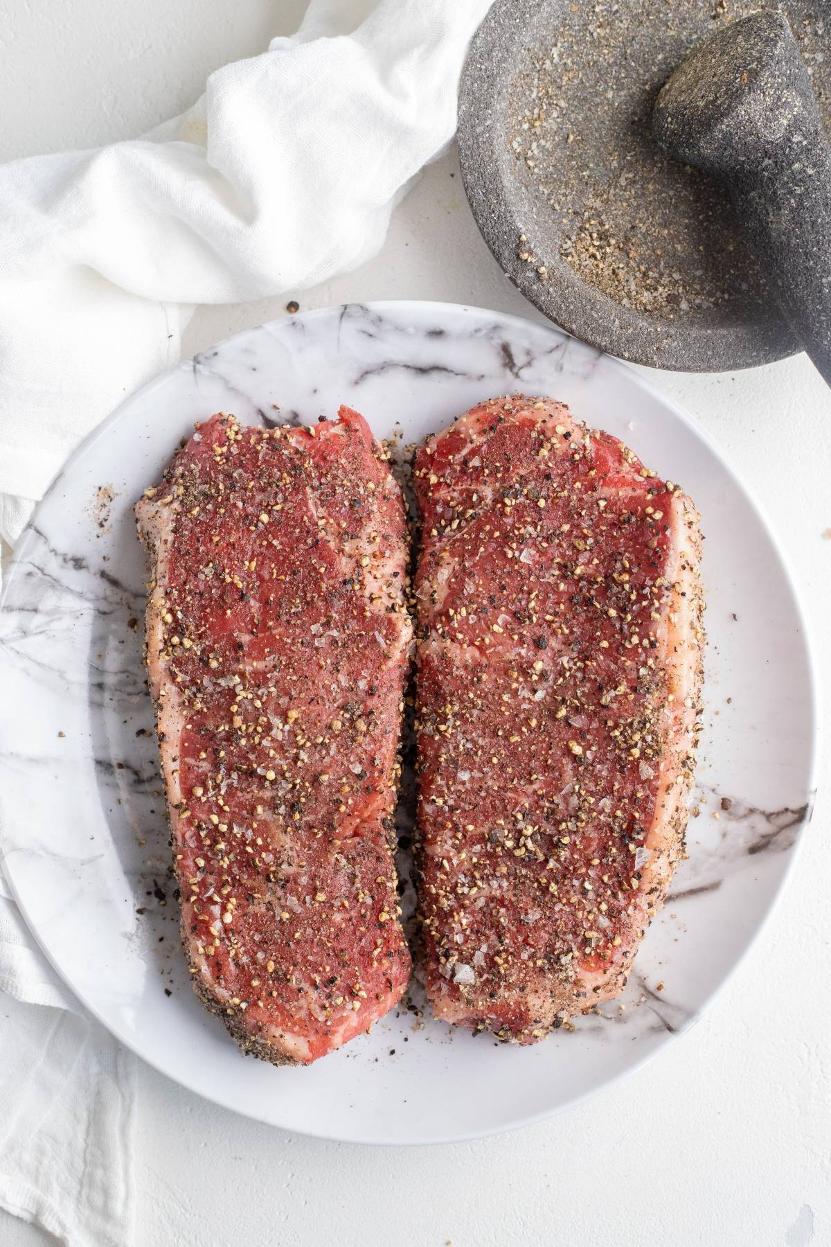 Two raw, seasoned steaks sit on a white plate with a mortar and pestle and a white cloth nearby—perfect for anyone looking to cook New York steak on a grill or searching for the ultimate BBQ steak recipe.
