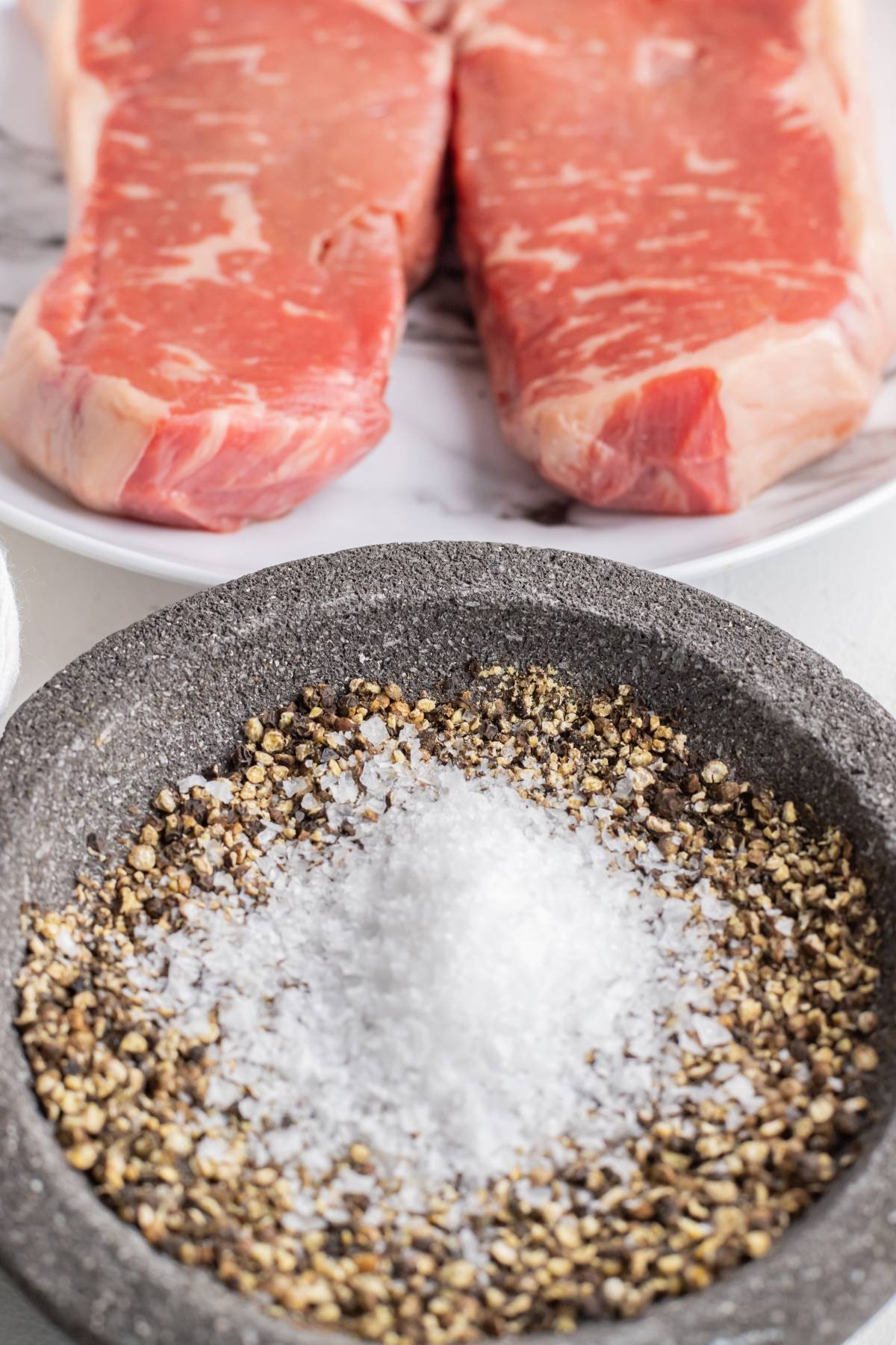 Two raw steaks on a plate behind a bowl of coarse salt and cracked black pepper, perfect for your favorite grilled steak topping or as the start of a classic BBQ steak recipe.
