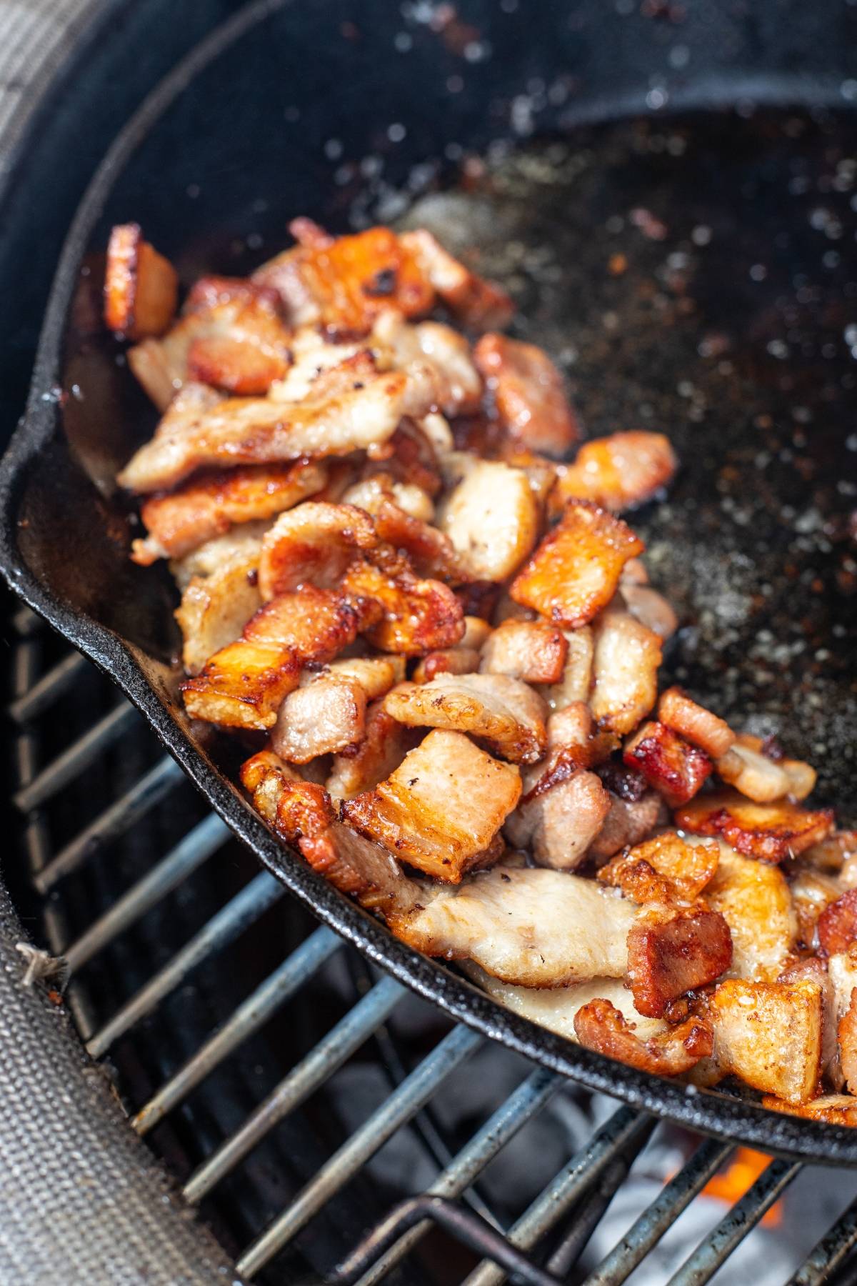 Crispy bacon pieces cooking in a cast iron skillet on a grill over an open flame—perfect as a grilled steak topping or to elevate your next BBQ steak recipe.