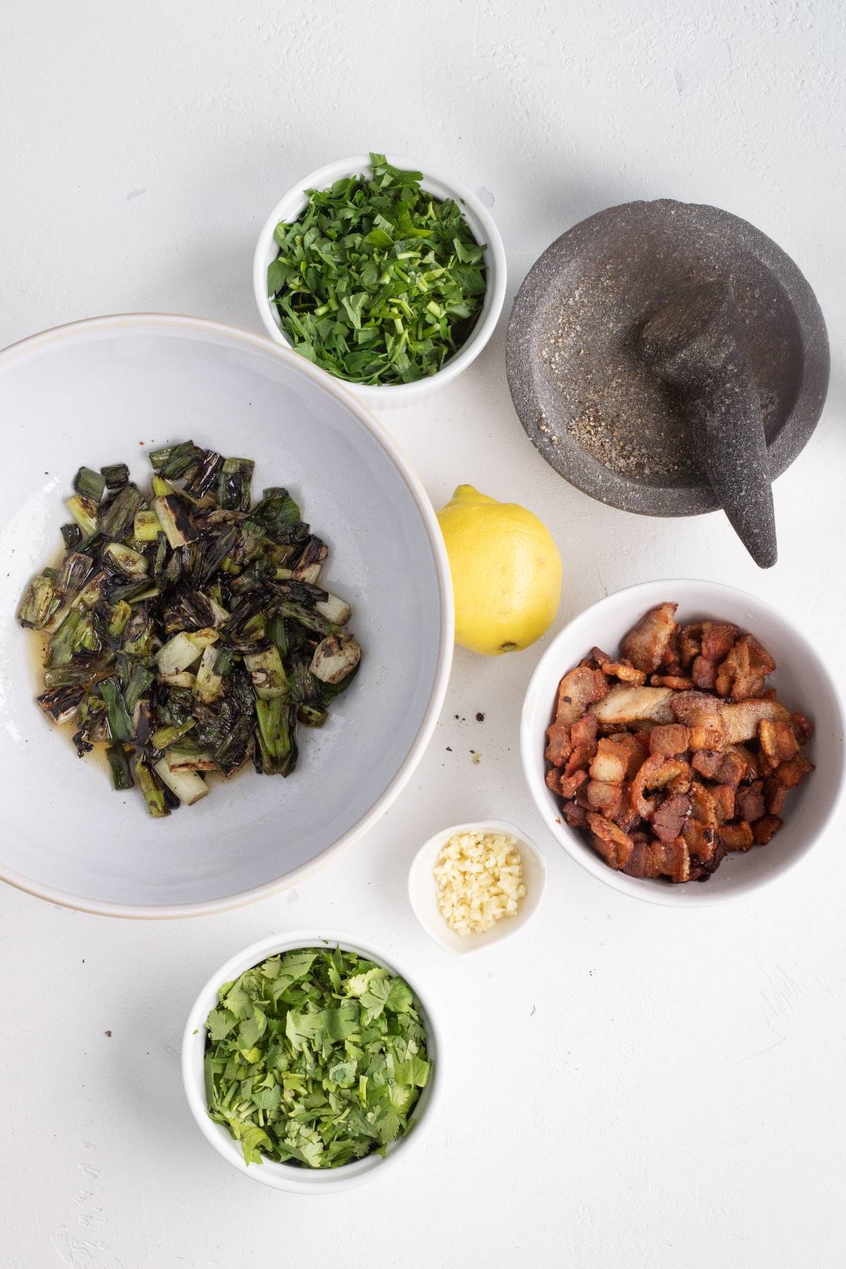 Bowl of grilled vegetables, bowls of herbs, bacon, minced garlic, a lemon, and a mortar and pestle on white surface—perfect accompaniments for your next BBQ steak recipe.
