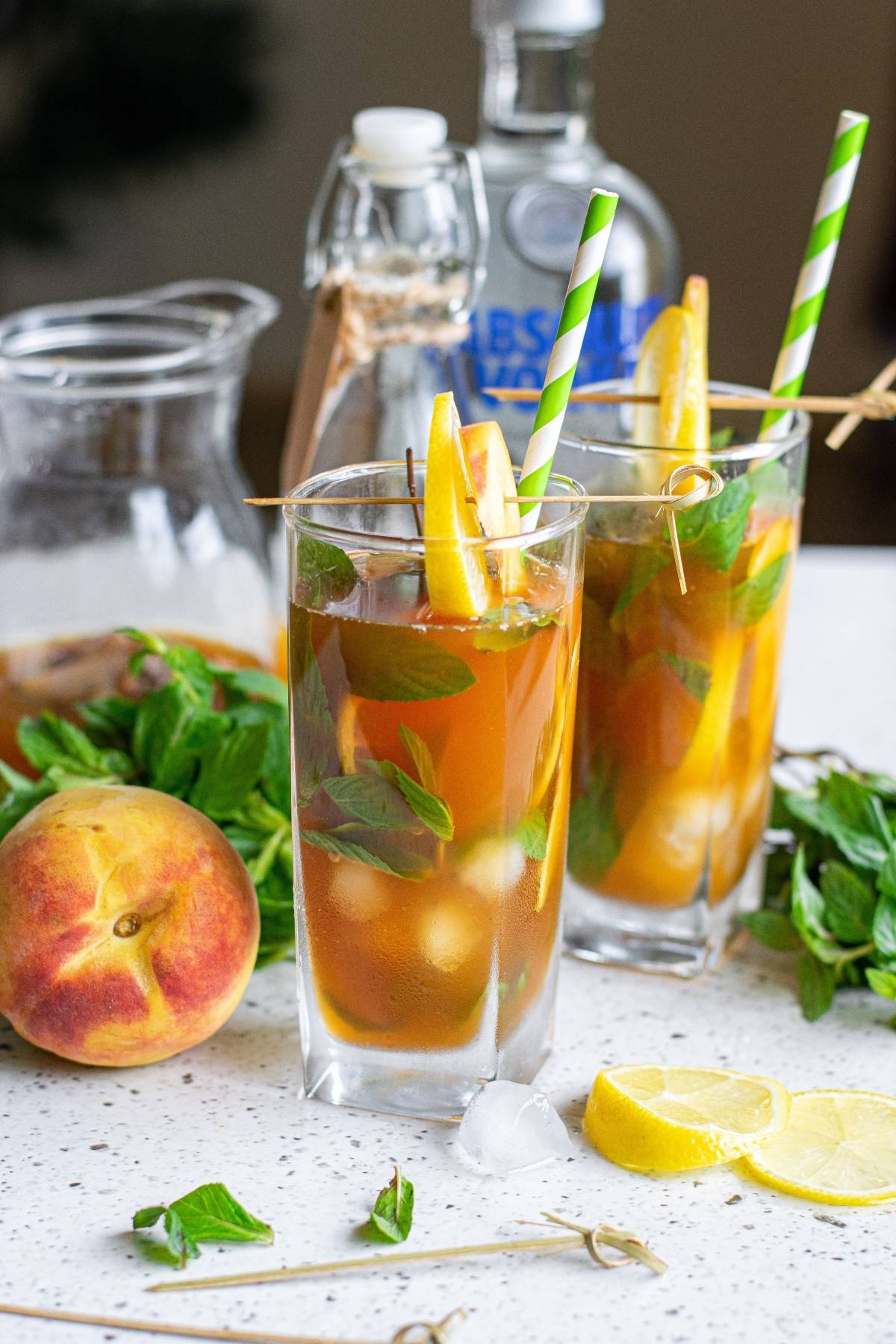 Two alcoholic peach tea cocktails with lemon, mint, and striped straws, surrounded by fresh peaches and lemon slices—a refreshing boozy iced tea recipe perfect for summer.