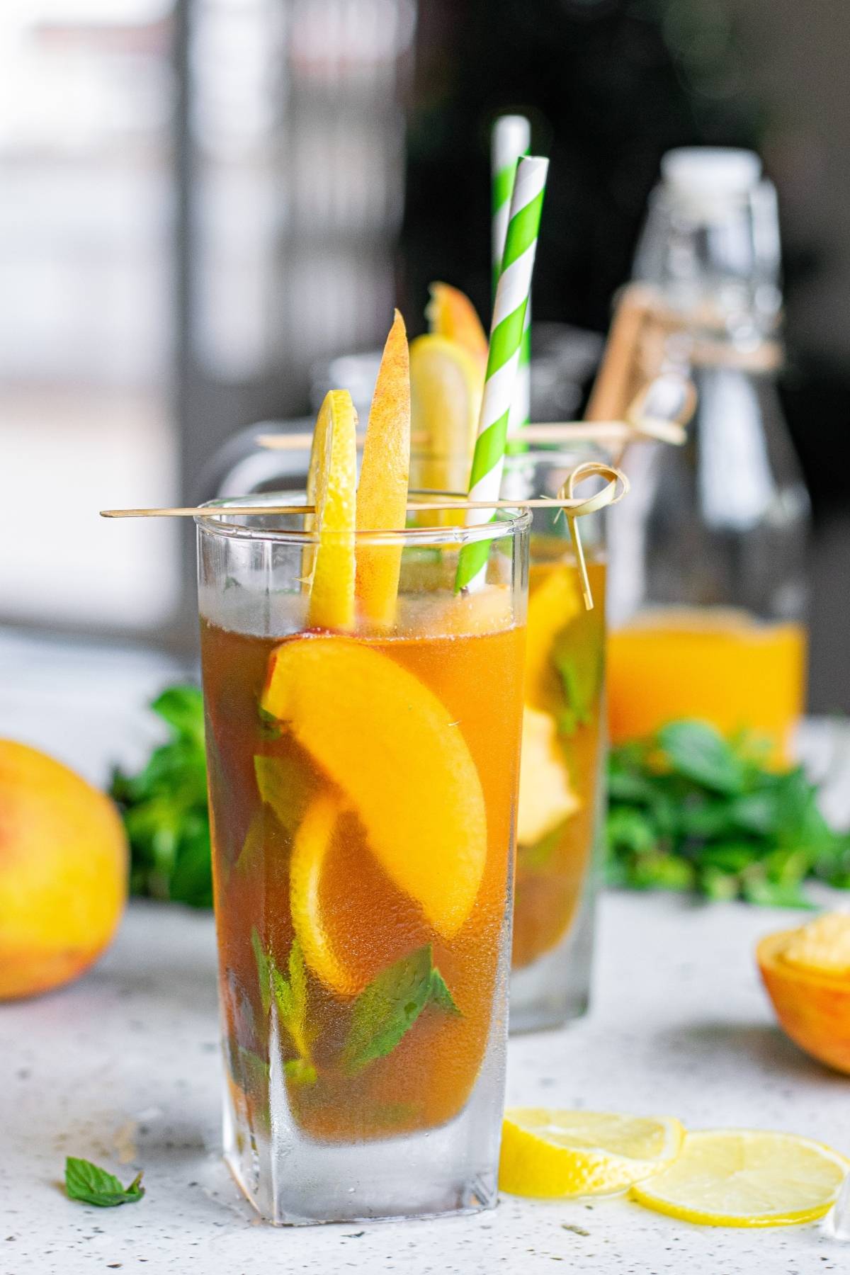 A tall glass of Hard Peach Iced Tea with peach slices, mint, lemon, and a green striped straw sits on the table, perfectly refreshing for summer.